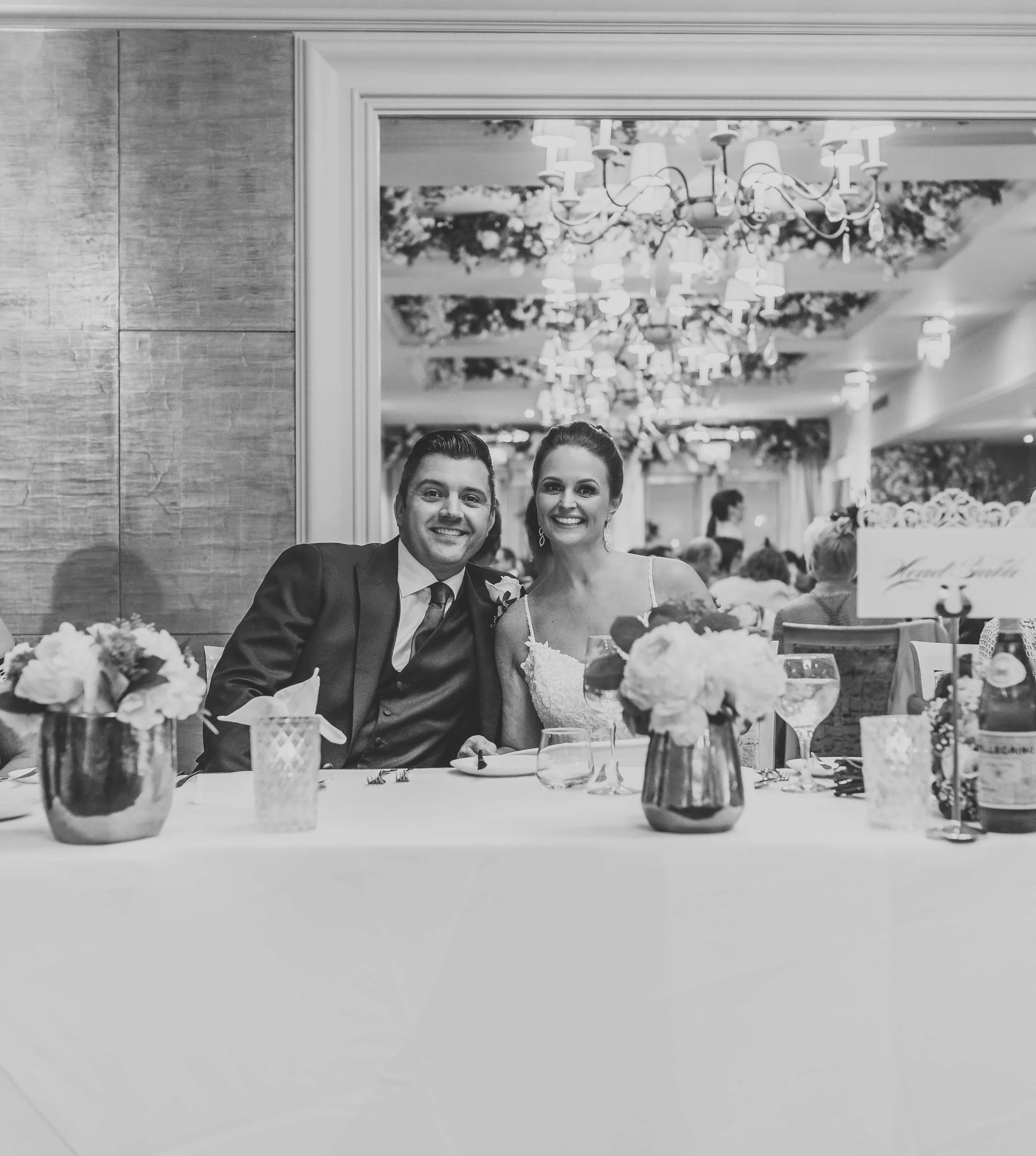 A black and white photograph of a smiling couple at a wedding reception, seated at a table with floral centerpieces, wine glasses, and tableware, with chandeliers and other guests in the background.