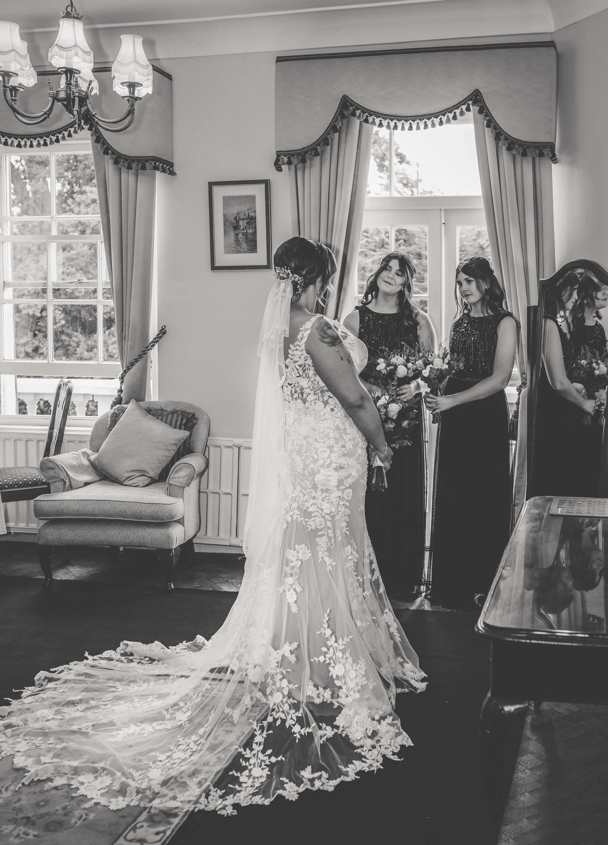 A bride in a lace wedding gown with a train and veil stands with her back to the camera, facing two women holding bouquets of flowers in a well-lit room with large windows, curtains, and a sofa.