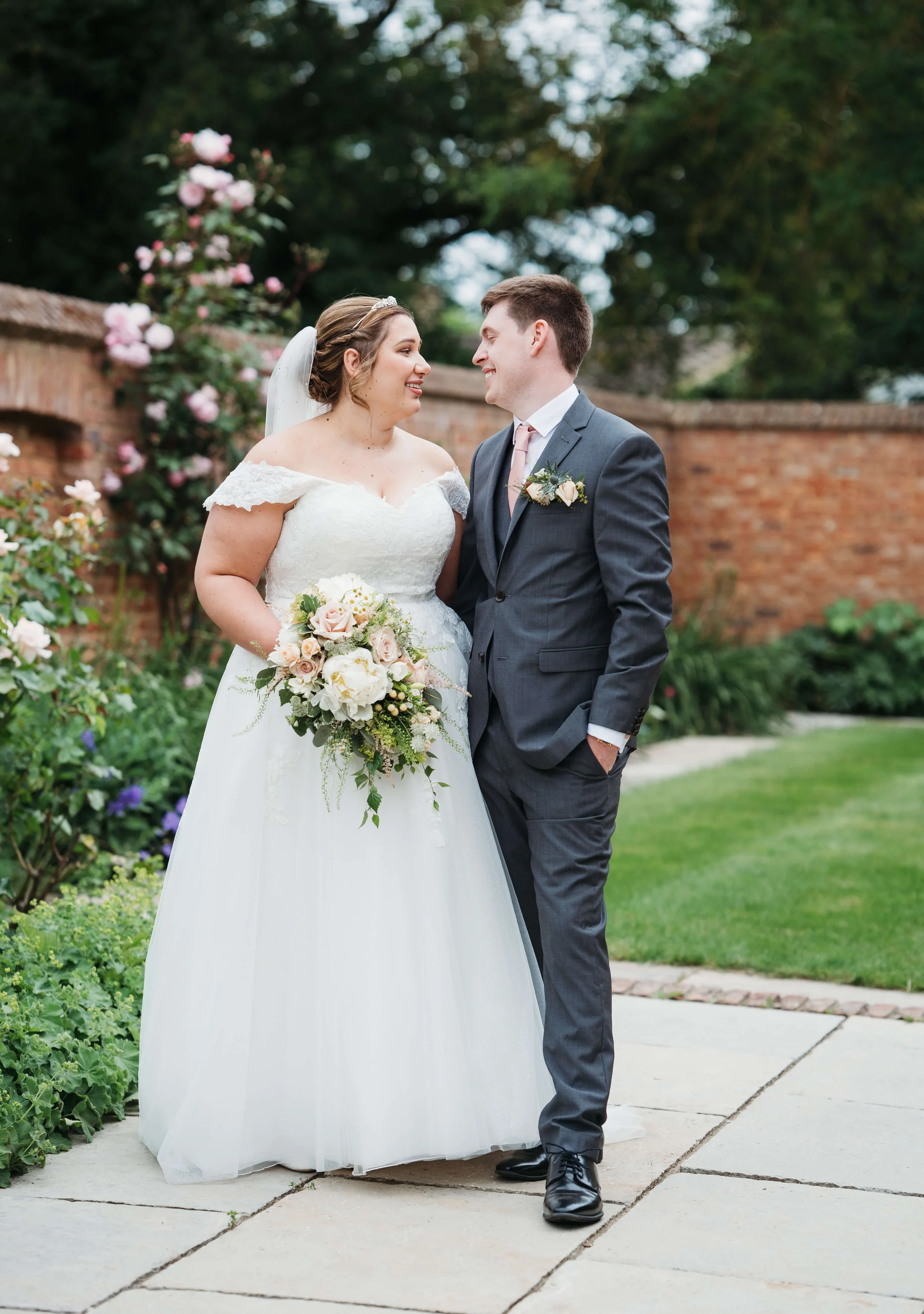 Bride and groom smiling at each other outdoors with a brick wall and greenery in the background.