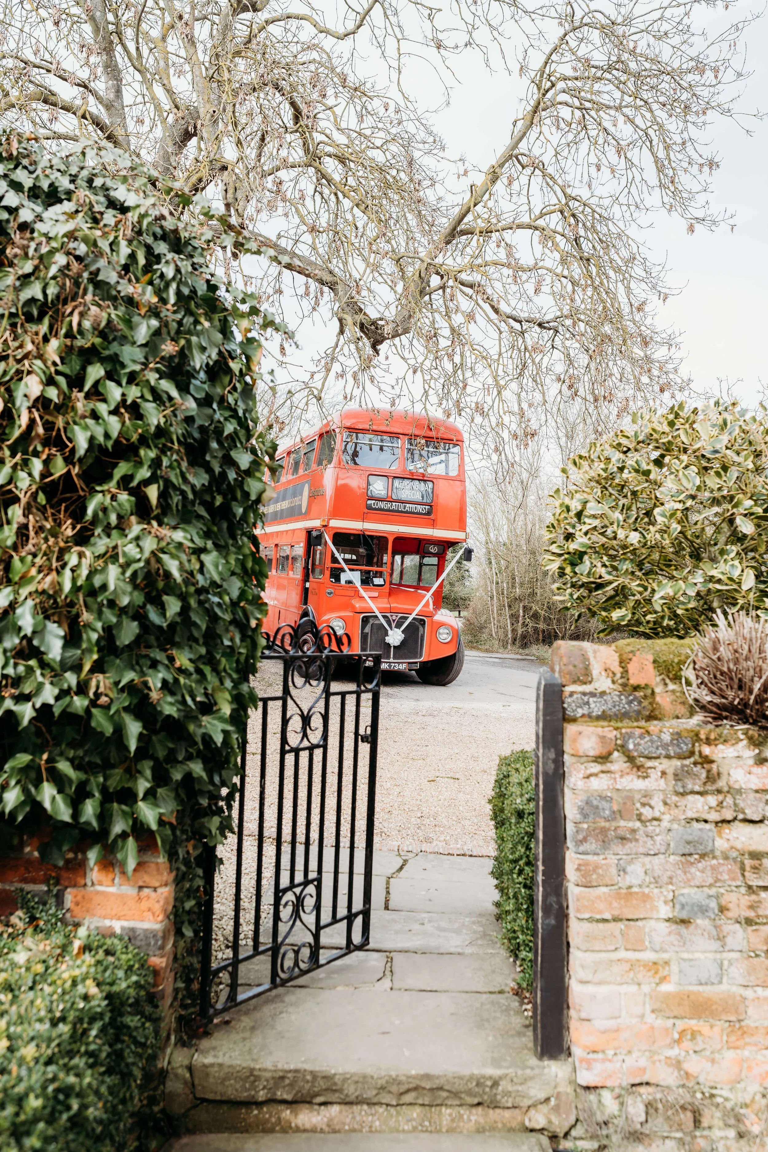 A vintage red double-decker bus parked on a street viewed through an open iron gate and garden hedge, with leafless trees in the background.