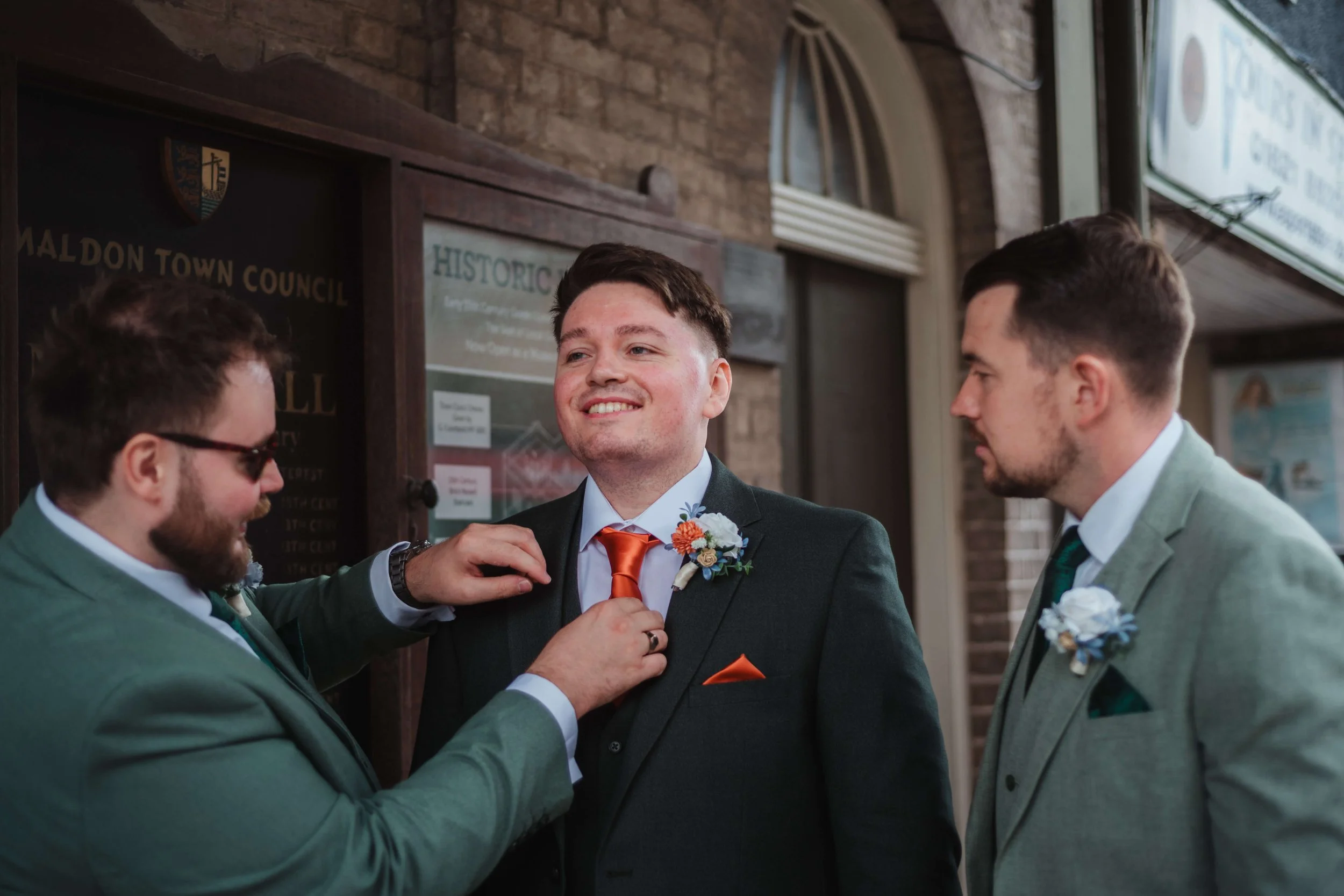 Three men in suits, with boutonnieres, at a wedding, with one adjusting the other's tie while another watches.
