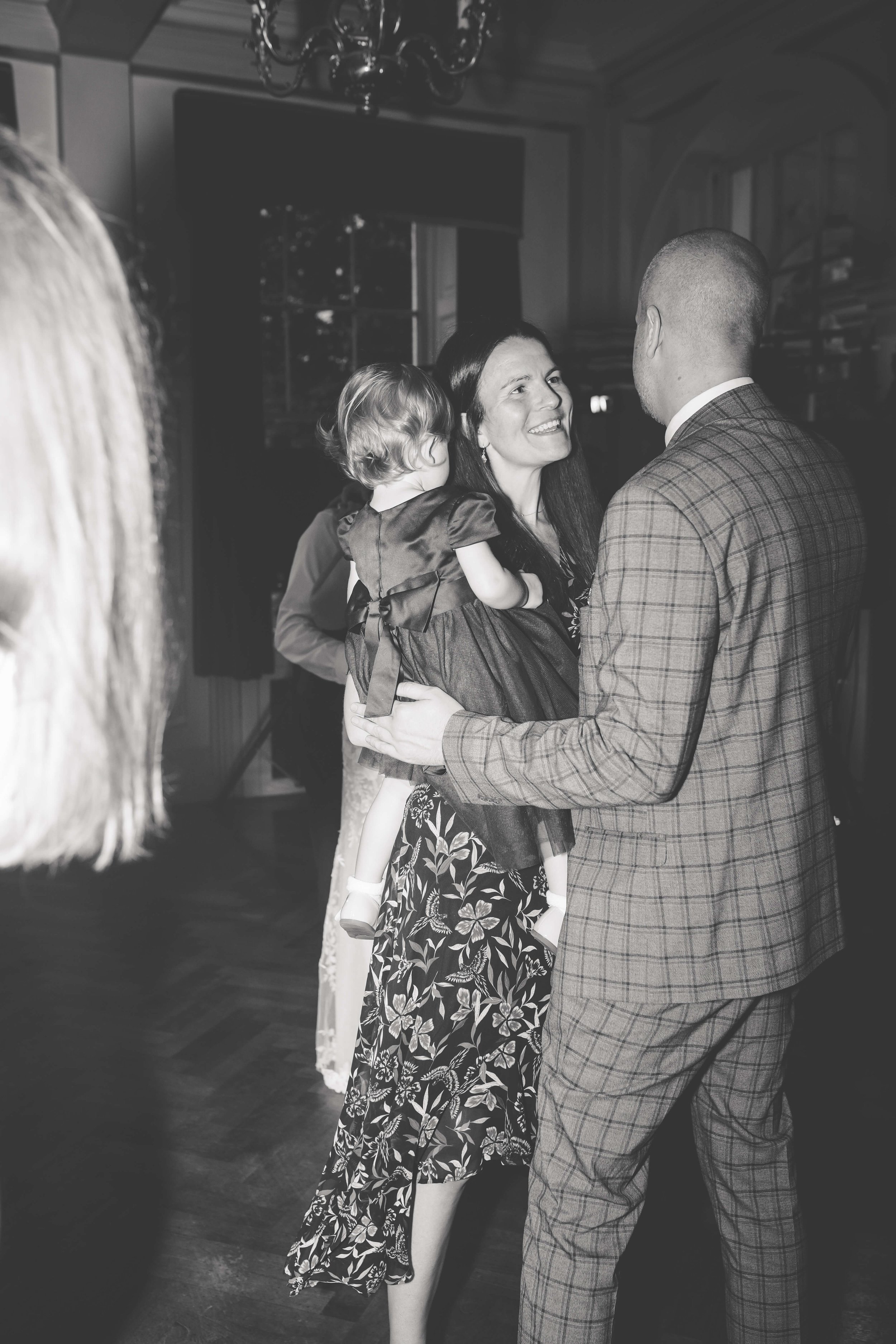 A woman and a man are dancing together at an indoor event, with the woman holding a young girl on her hip. The woman is smiling and looking at the man, while the girl is facing away. The image is in black and white.