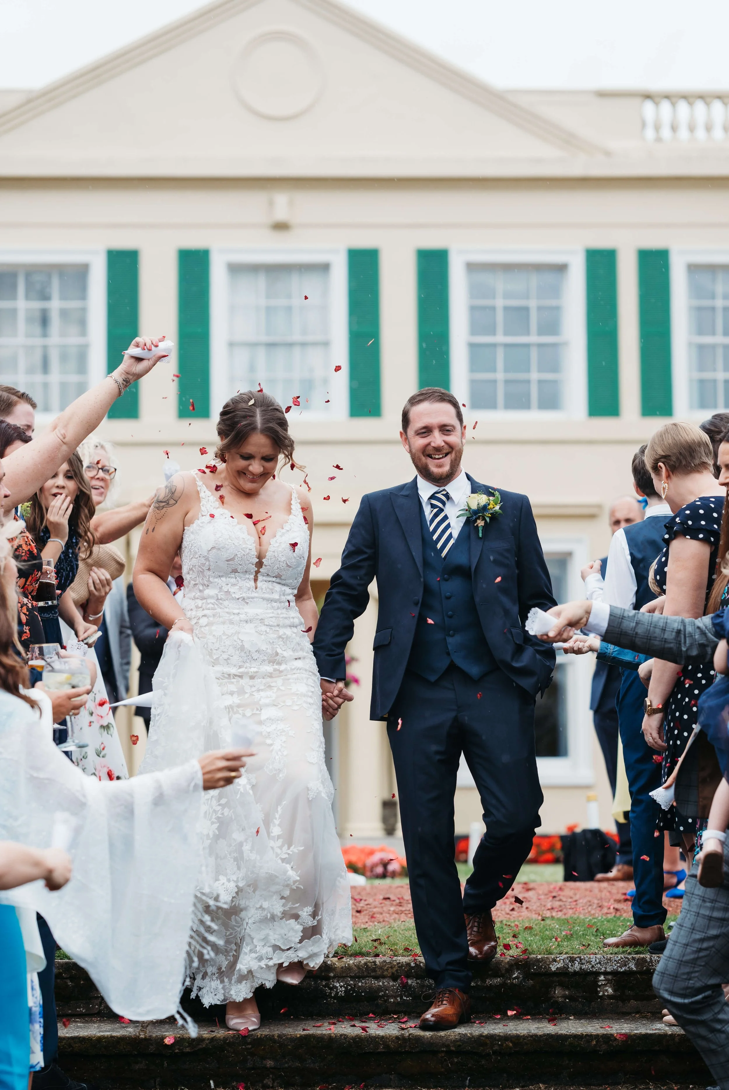 Happy newlywed couple walking hand in hand outside a house, surrounded by friends throwing flower petals.