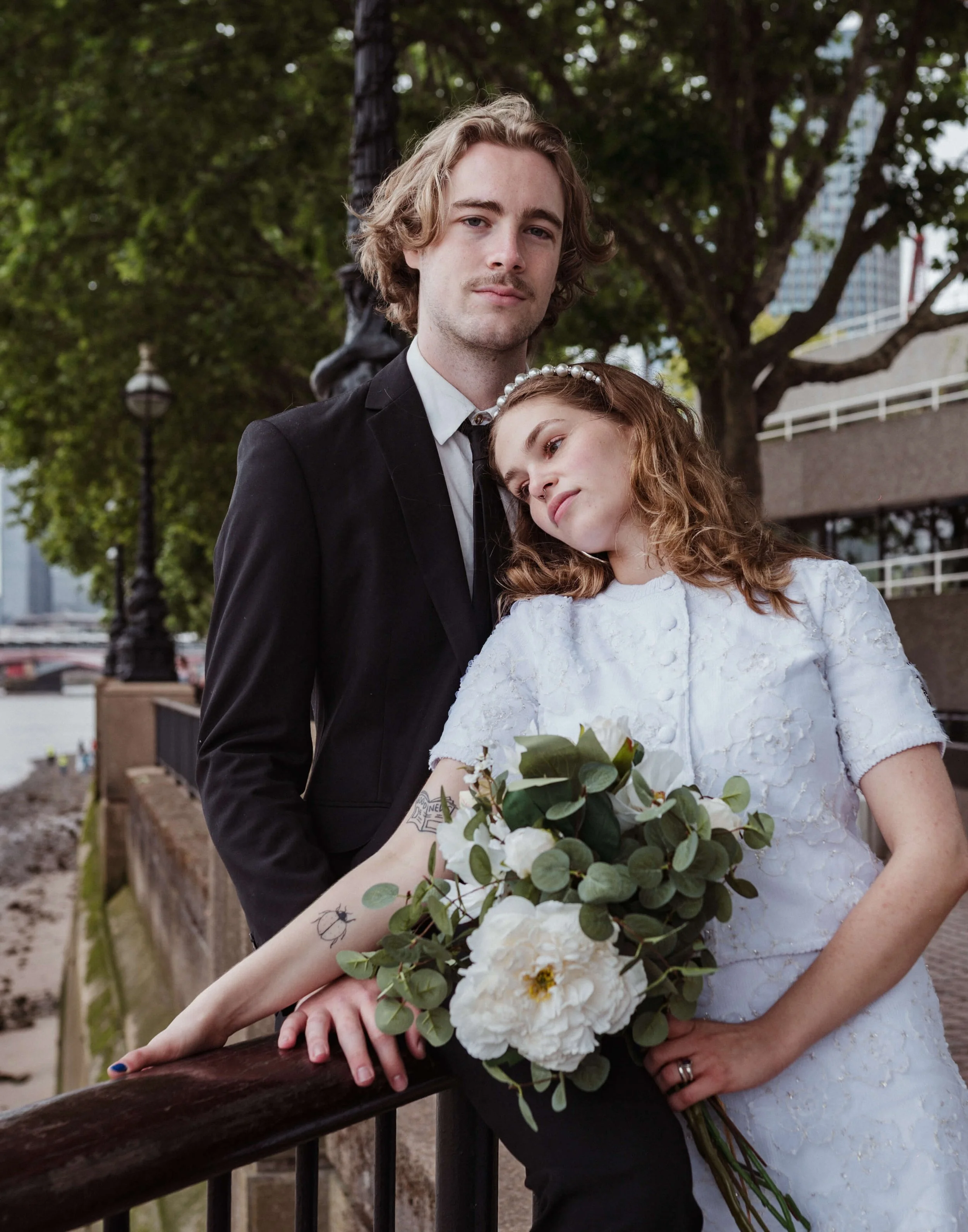 A couple dressed in wedding attire stands outdoors near a river, with city buildings and trees in the background. The woman is holding a bouquet of white flowers and greenery, and the man is wearing a black suit with a white shirt.