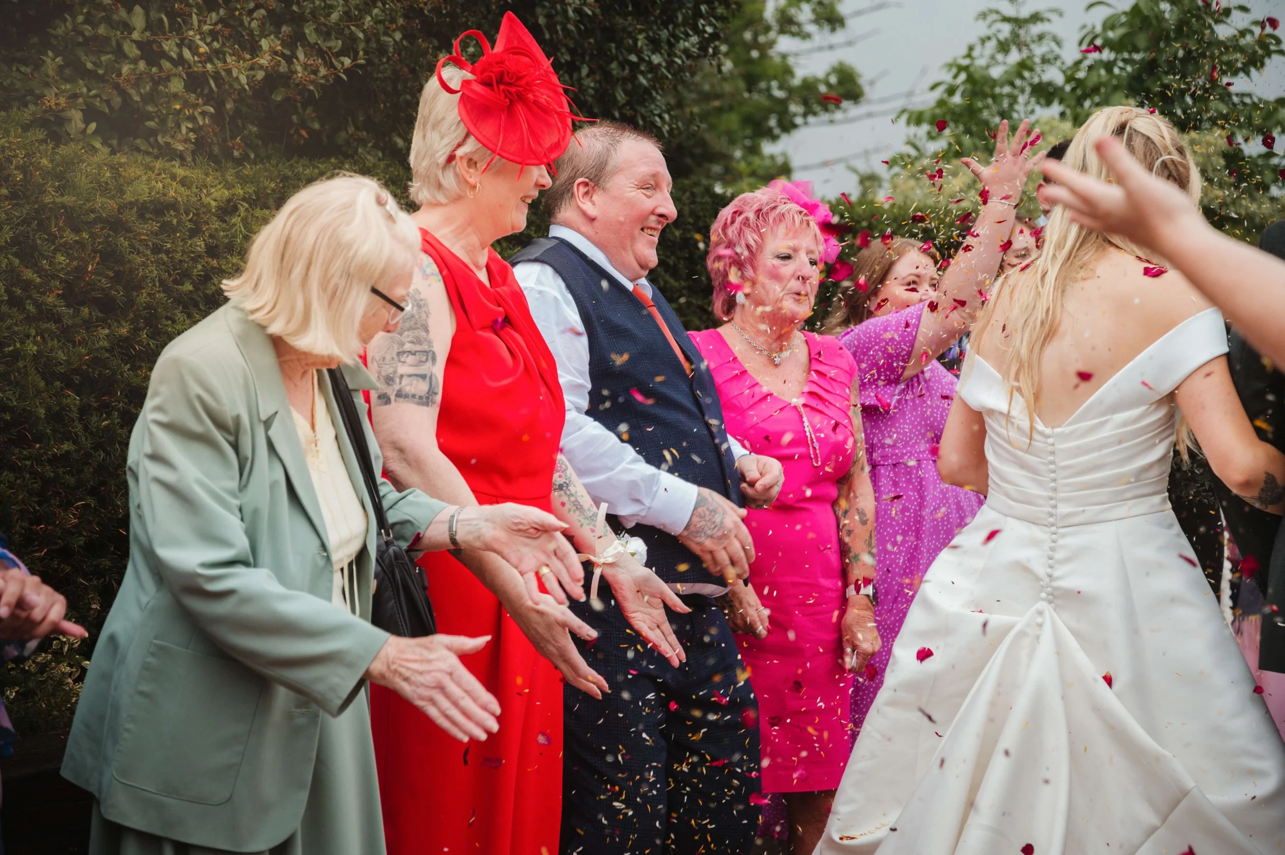 Group of people celebrating at a wedding, with confetti in the air, standing outdoors with greenery in the background.