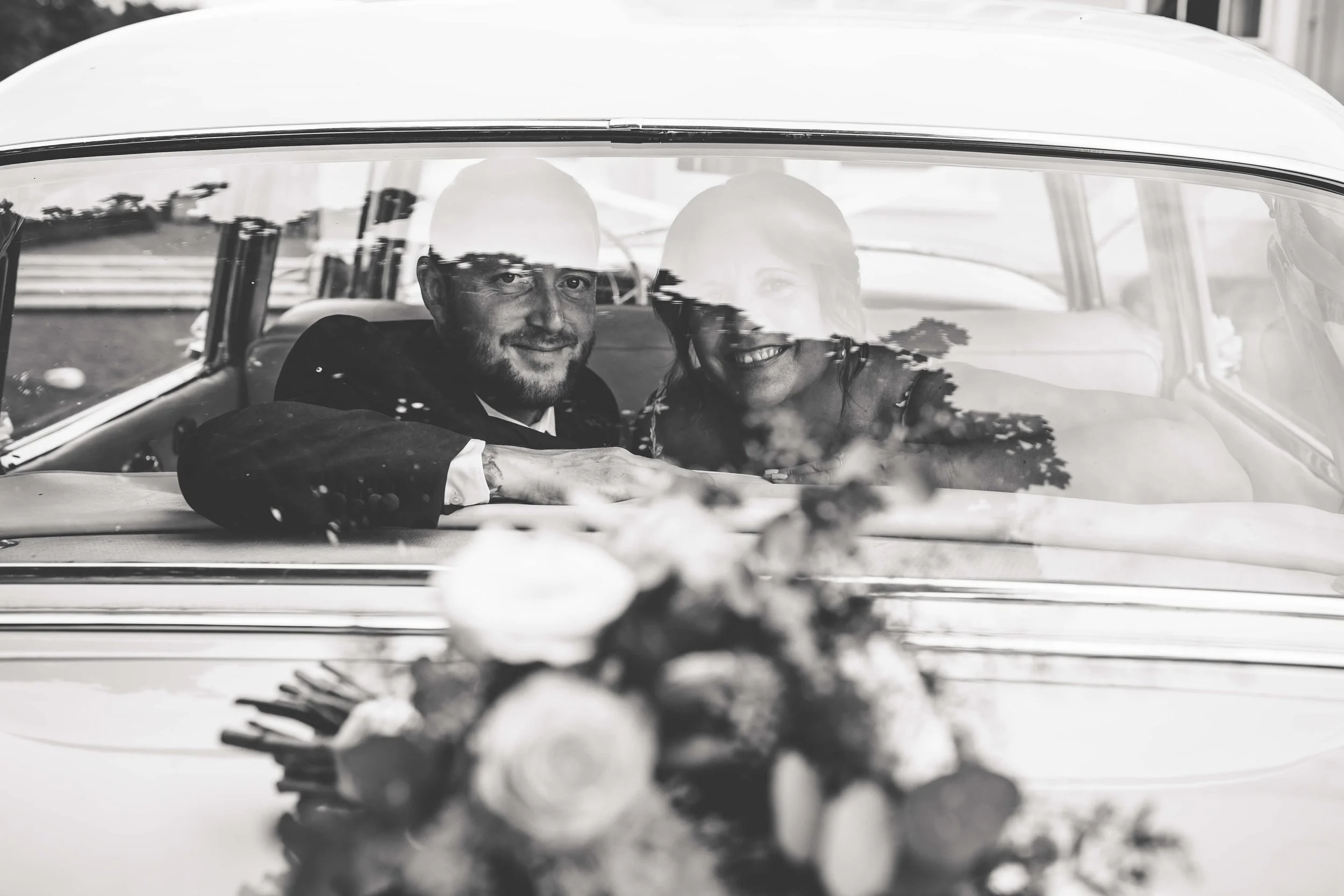 A black and white photo of a smiling bride and groom sitting in a vintage car, with a bouquet placed on the hood in the foreground.