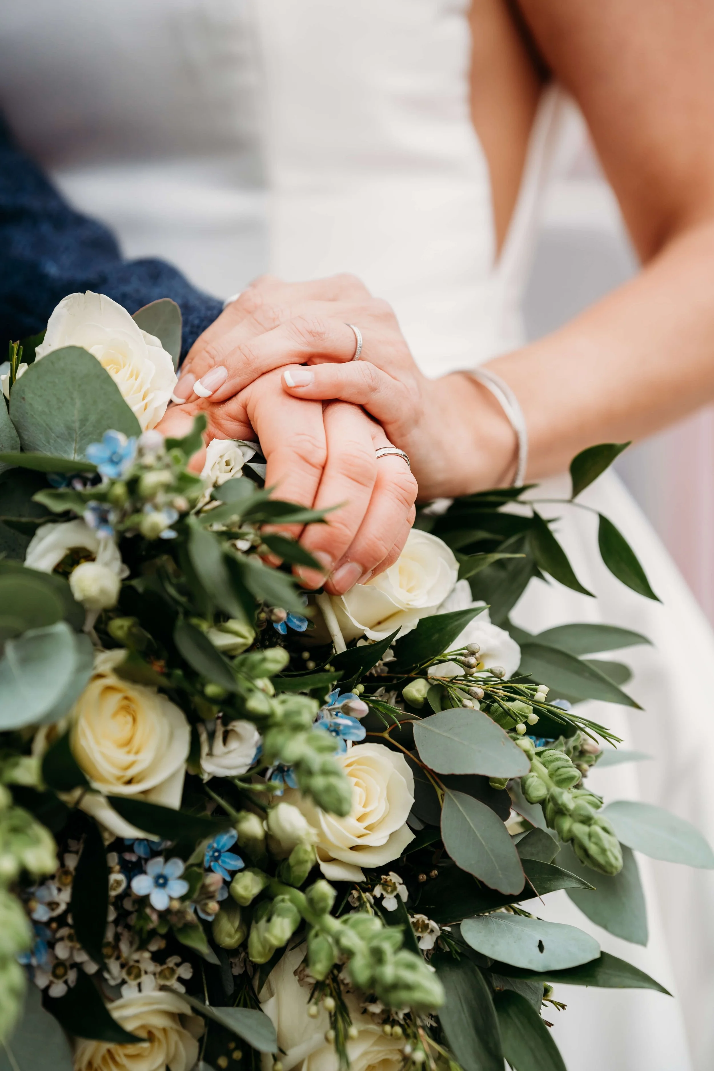Close-up of two hands holding each other, one with a wedding ring. They are near a bouquet of white roses and greenery.