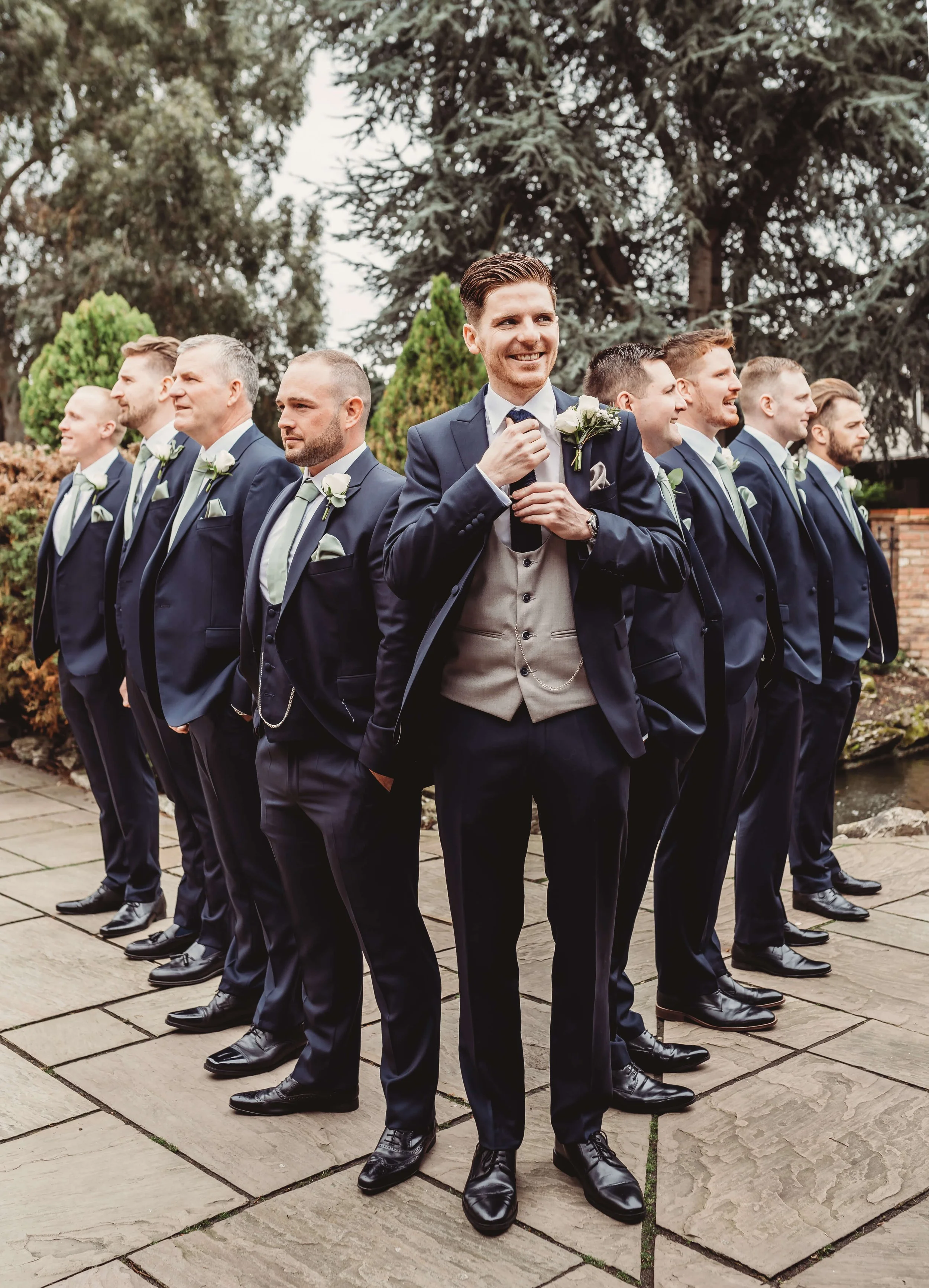 Group of groomsmen in formal suits, likely at a wedding, standing outdoors on a stone pavement with trees in the background. The man at the front is smiling and adjusting his tie.