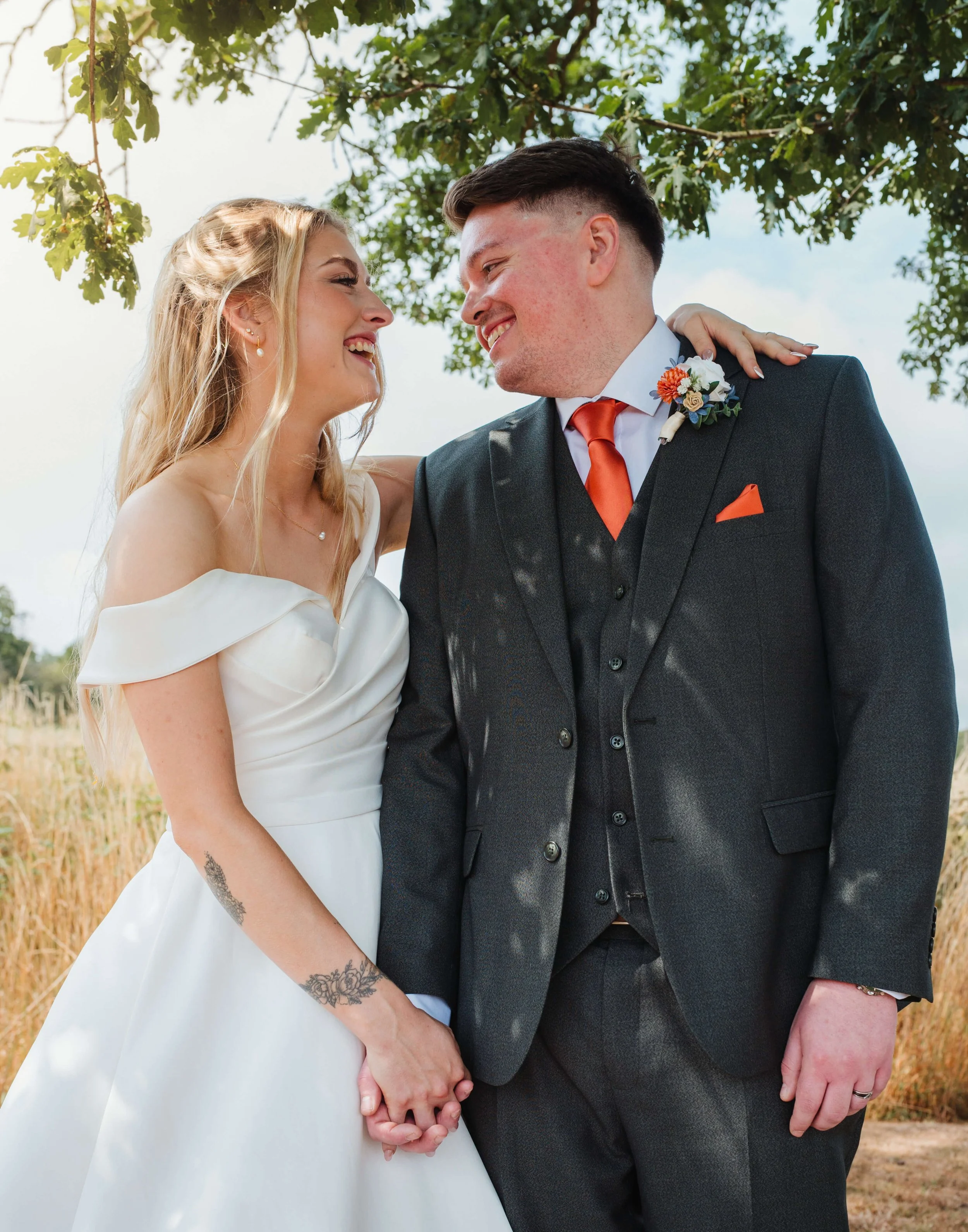 A bride and groom smiling and holding hands, standing outdoors under a tree on their wedding day.