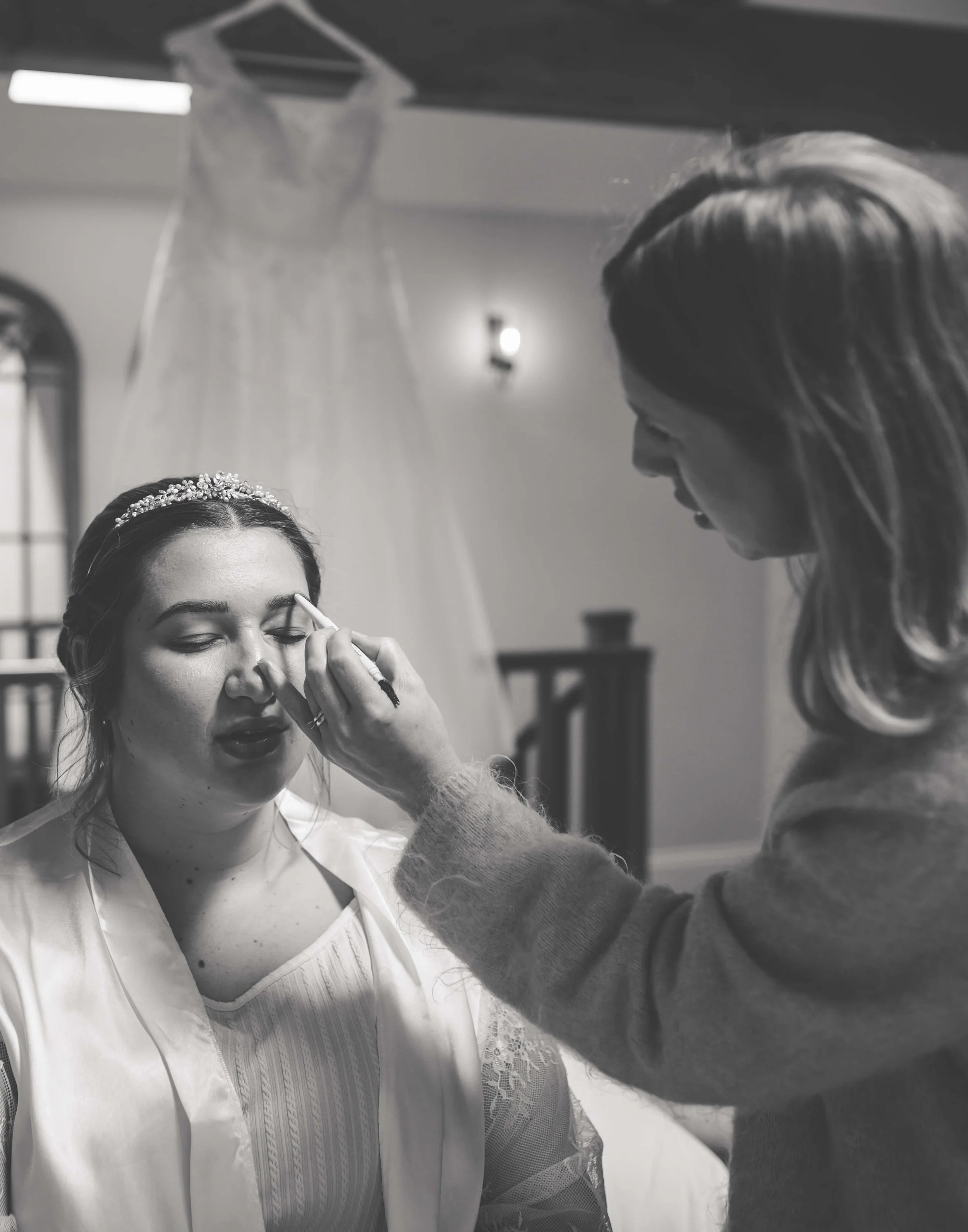A woman is getting her makeup done by a makeup artist in a room, with a wedding dress hanging in the background.