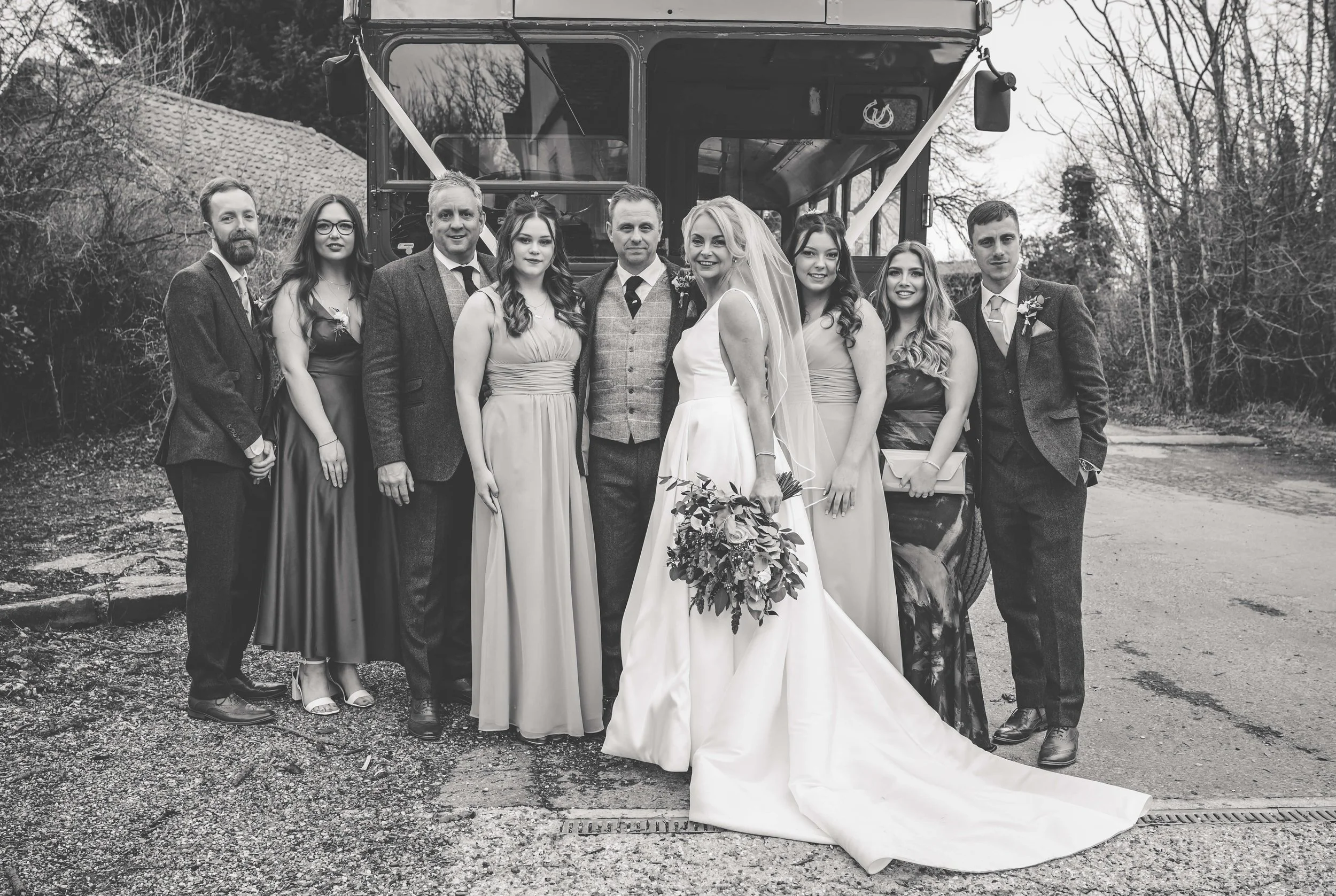Black and white photo of a wedding group standing outdoors in front of a tractor. The bride in a white gown and veil holds a bouquet of flowers, surrounded by bridesmaids and groomsmen dressed in formal attire.
