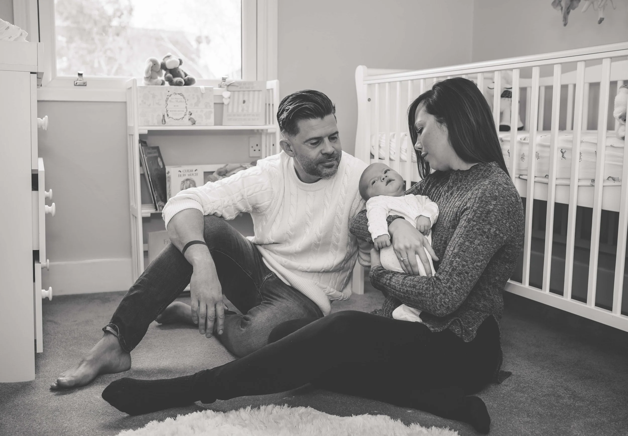 A family sitting on the floor of a nursery, with a man, woman, and baby. The woman holds the baby, and the man looks at the baby and woman. The nursery has a crib, books, and stuffed animals.