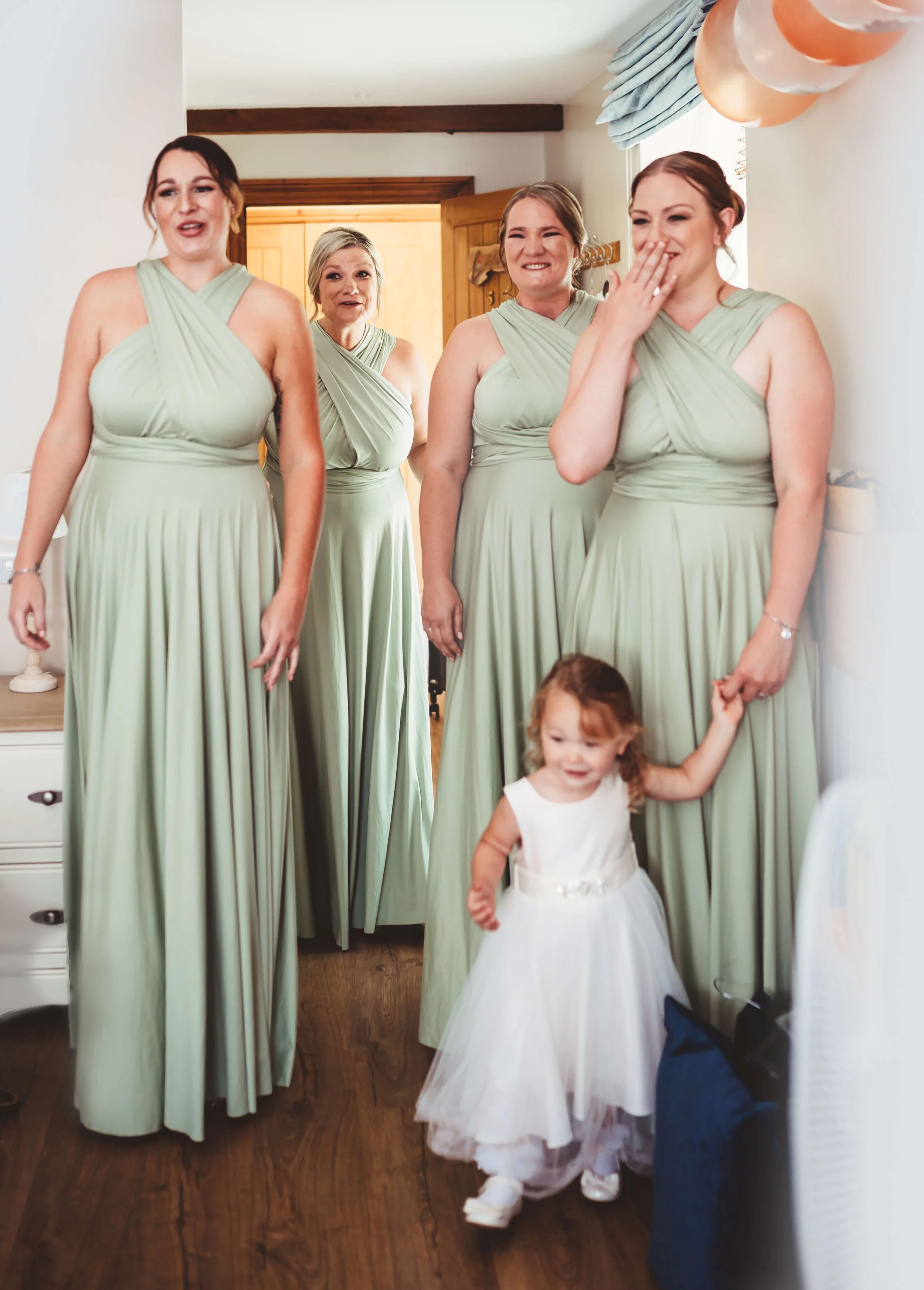 Four women and a young girl in a room, the women are wearing matching sage green dresses, and the girl is in a white dress. The women are smiling and one woman is covering her mouth with her hand, reacting to an event or joke. The room has balloons a