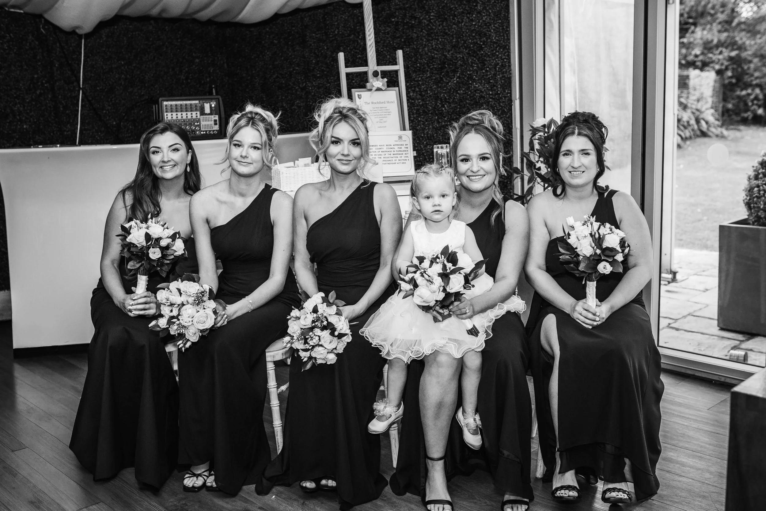 Six women and one young girl sitting on chairs at a wedding, holding bouquets, indoors with a sliding glass door and garden visible in the background.
