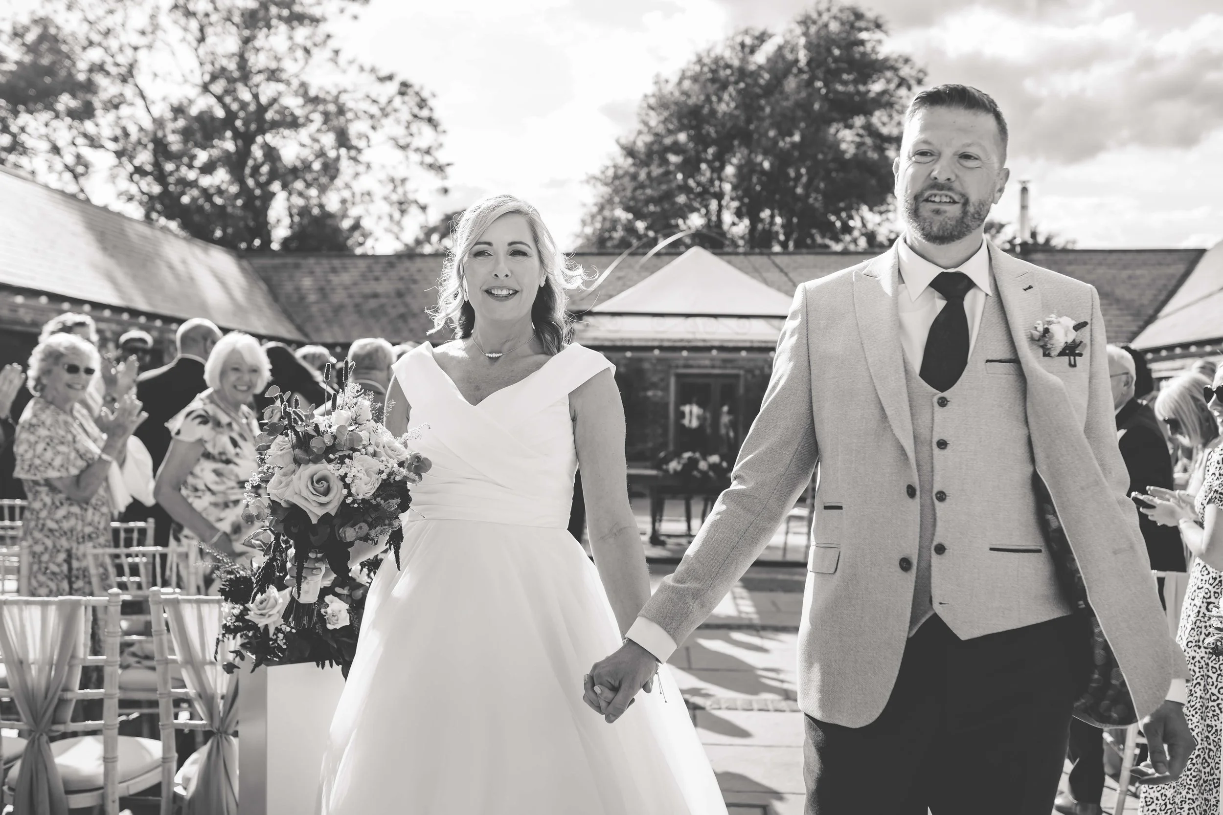 A bride and groom holding hands and walking outdoors at a wedding ceremony, with guests clapping in the background.