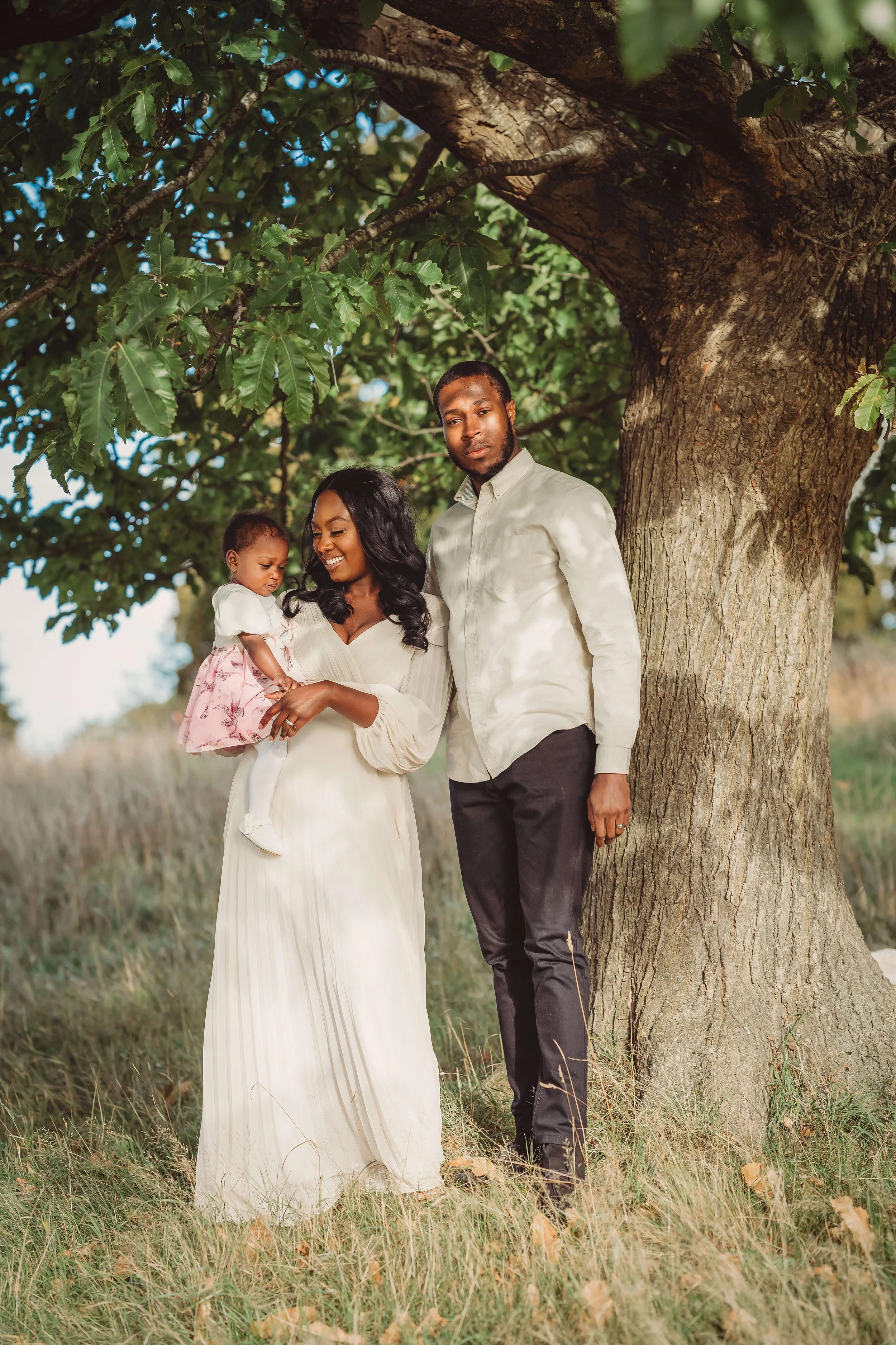 A family of three standing outdoors under a large tree, with the mother holding their young daughter and the father standing beside them, all dressed in light-colored clothing.