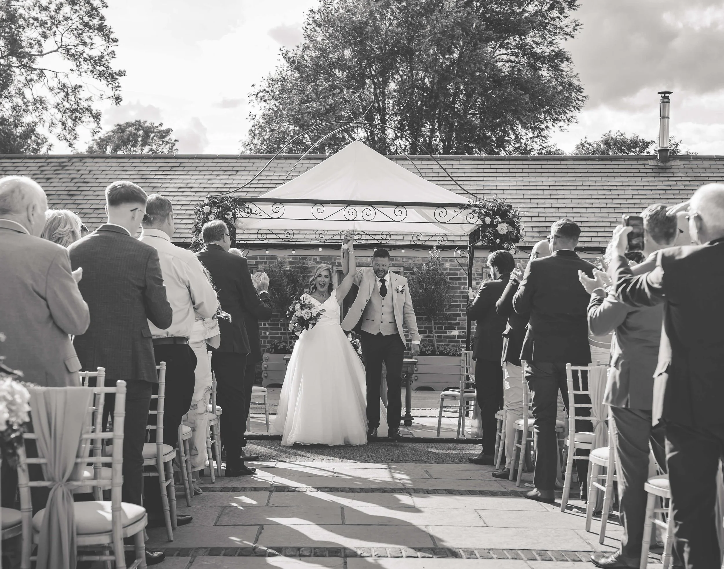 A black and white photo of a wedding ceremony outdoors with a bride and groom holding hands and raising their clasped hands in celebration. The bride is holding a bouquet of flowers, both are smiling. Friends and family are standing and clapping on e