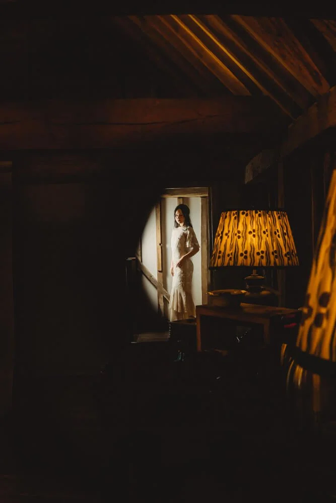 A woman in a white lace dress standing in a doorway, illuminated by natural light, with a lamp and wooden interior in the foreground.
