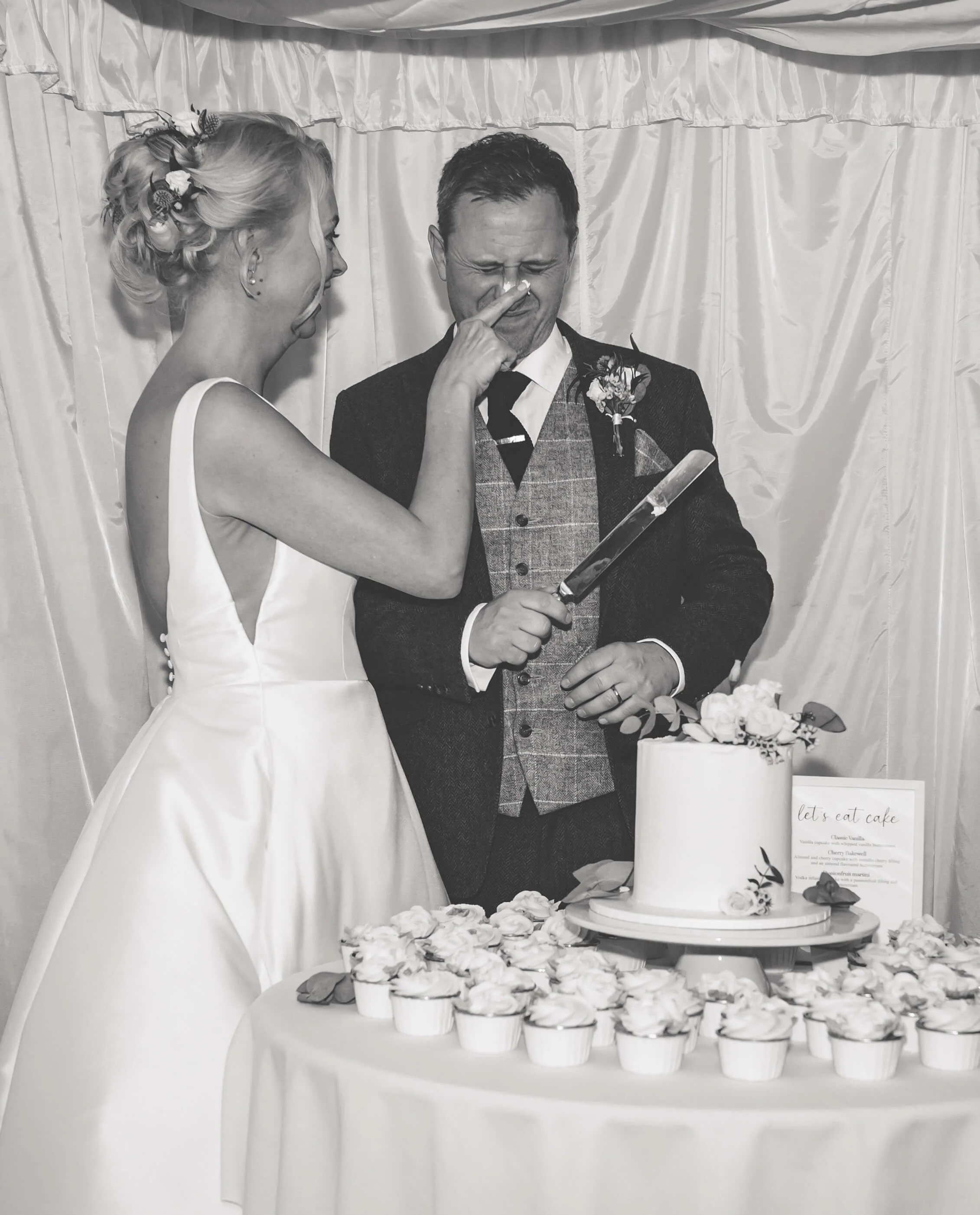 Bride and groom at wedding reception, the bride playfully touching the groom's nose with her finger. There is a wedding cake and cupcakes on the table in front of them.