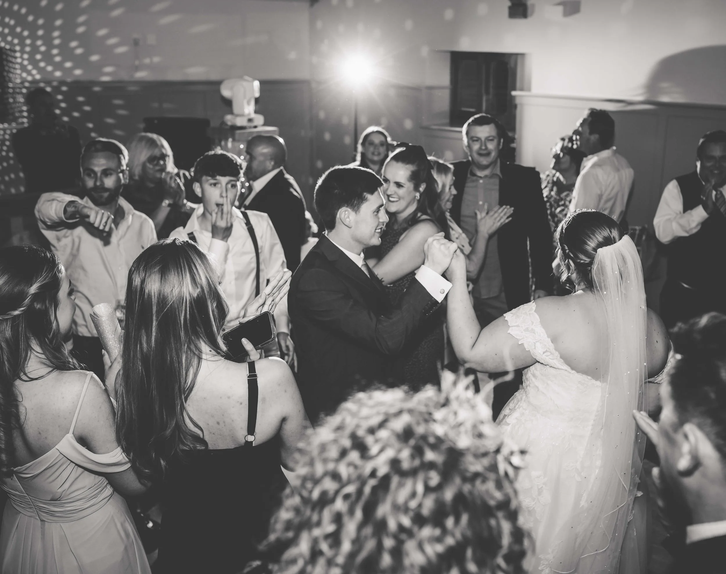 A black-and-white photo of a wedding reception dance floor with people dancing and celebrating, featuring a bride and groom sharing a dance