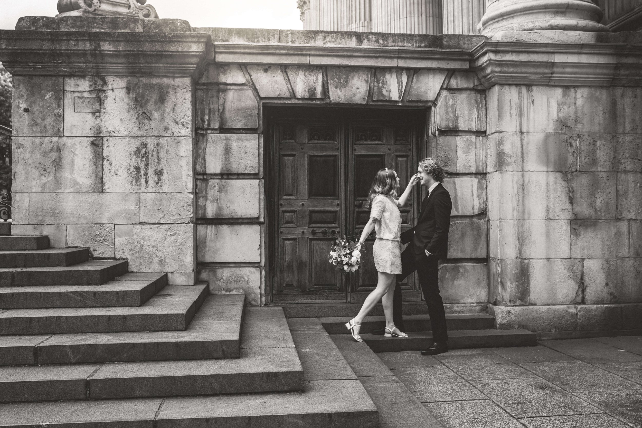 A black and white photo of a couple in wedding attire standing in front of a large stone door on a set of stairs, with the woman holding a bouquet of flowers and touching the man's face.