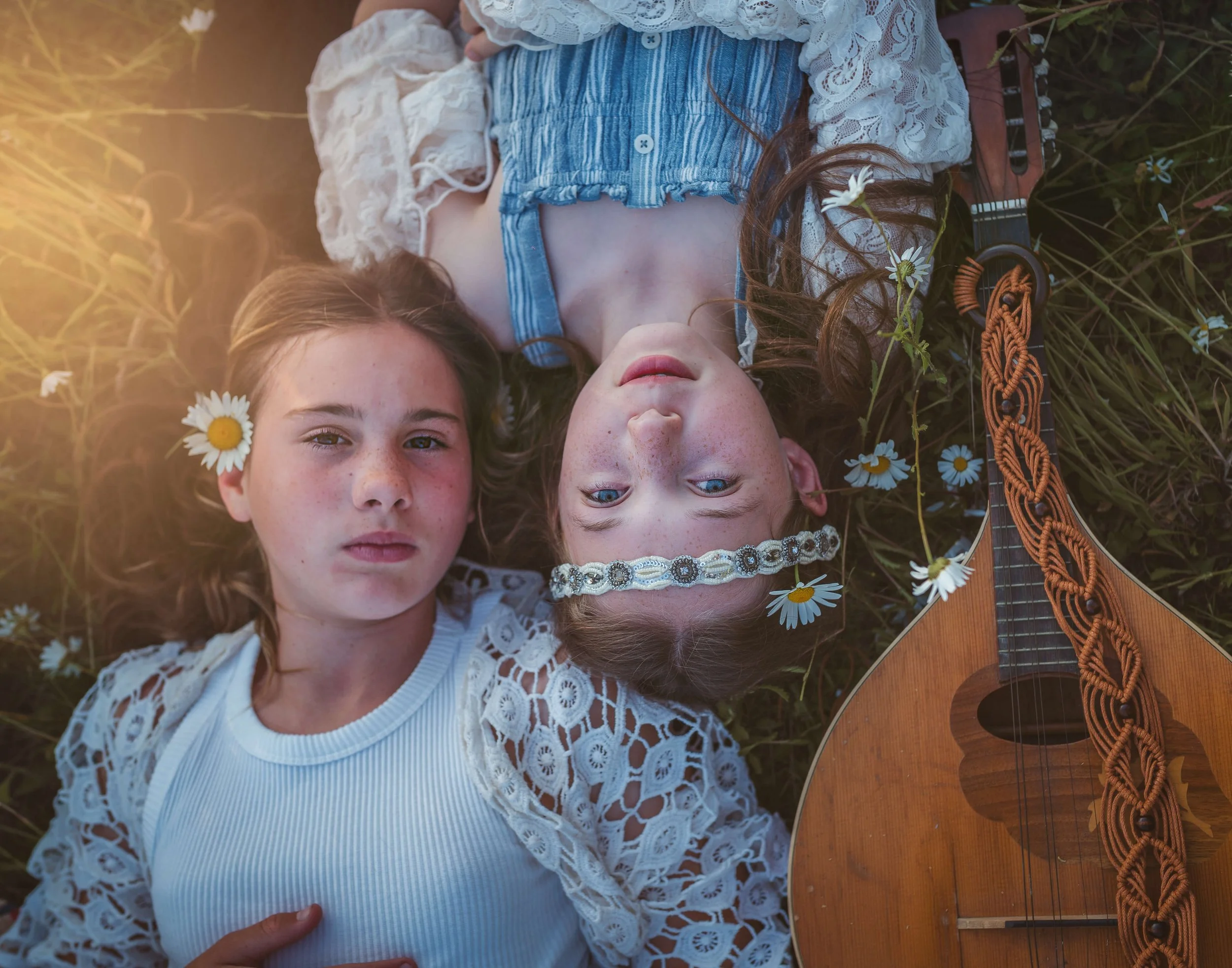 Two young girls lying on grass among daisies, one with a daisy in her hair, one with a flower headband, and a guitar beside them.