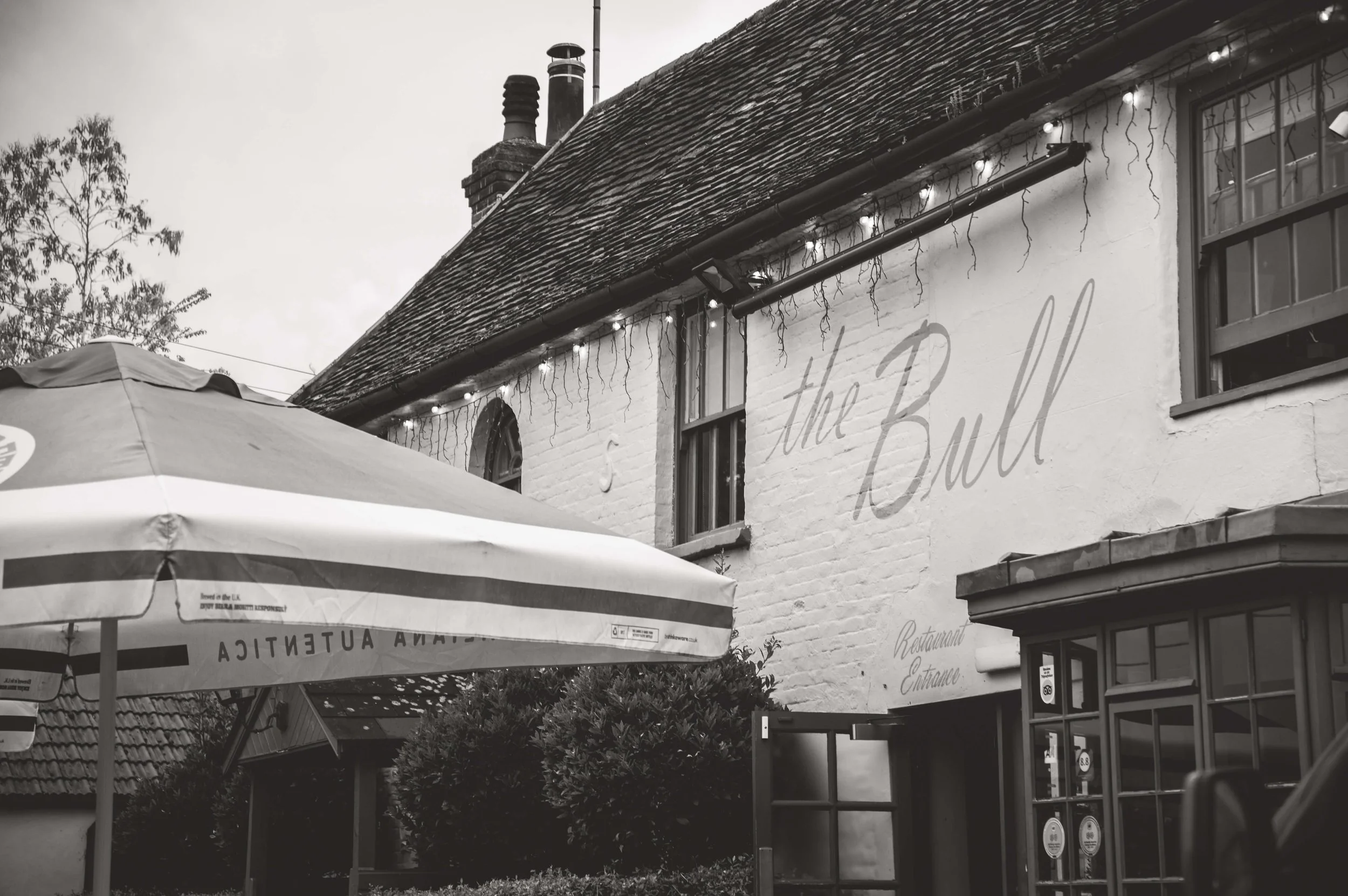 Exterior of a building with a large sign that reads 'the Bull,' decorated with string lights, and an outdoor umbrella partially visible in the foreground.