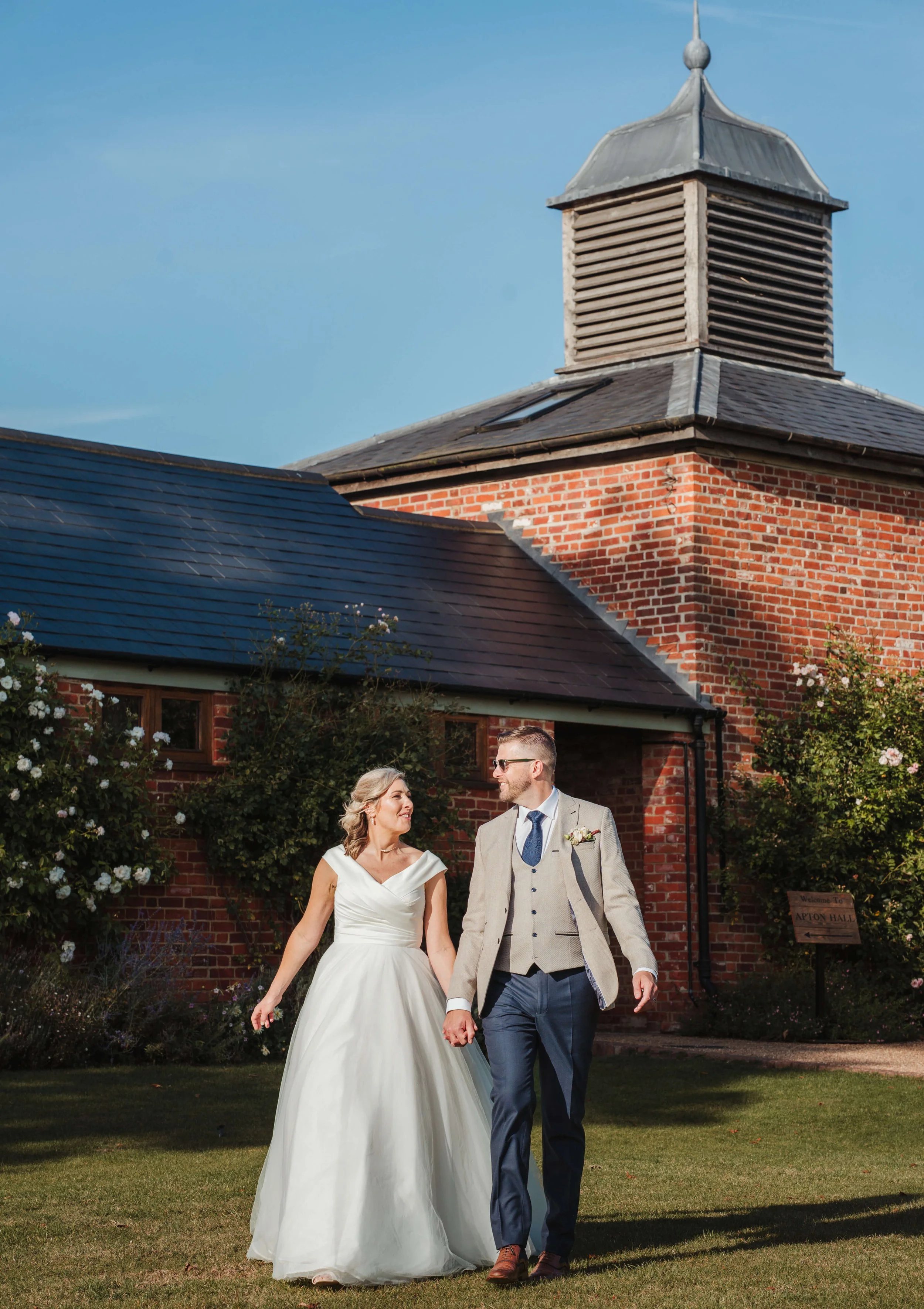 A bride and groom walking hand in hand outside a red brick building with a small tower and dark roof, surrounded by greenery and blooming flowers.