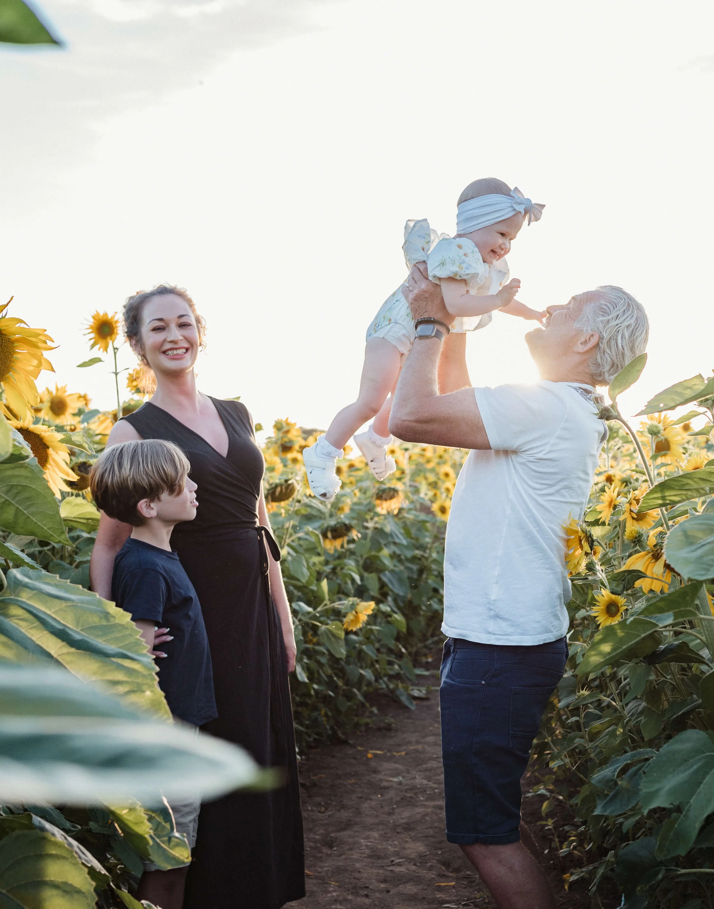 Family enjoying a sunflower field during sunset, with a man lifting a young girl, a woman and a boy looking on
