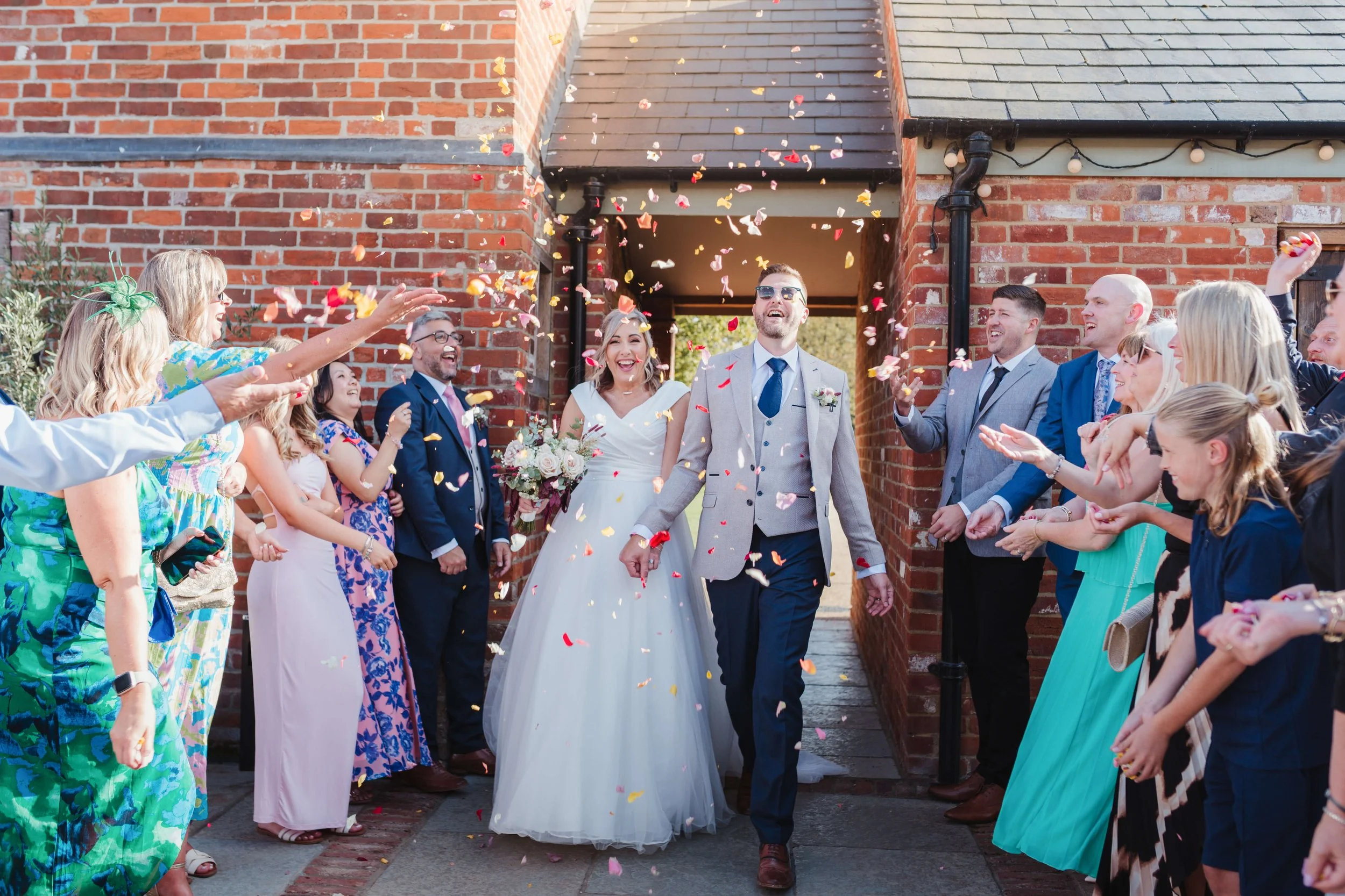 A bride and groom walking through a crowd of friends and family throwing flower petals during their wedding celebration outside a brick building.