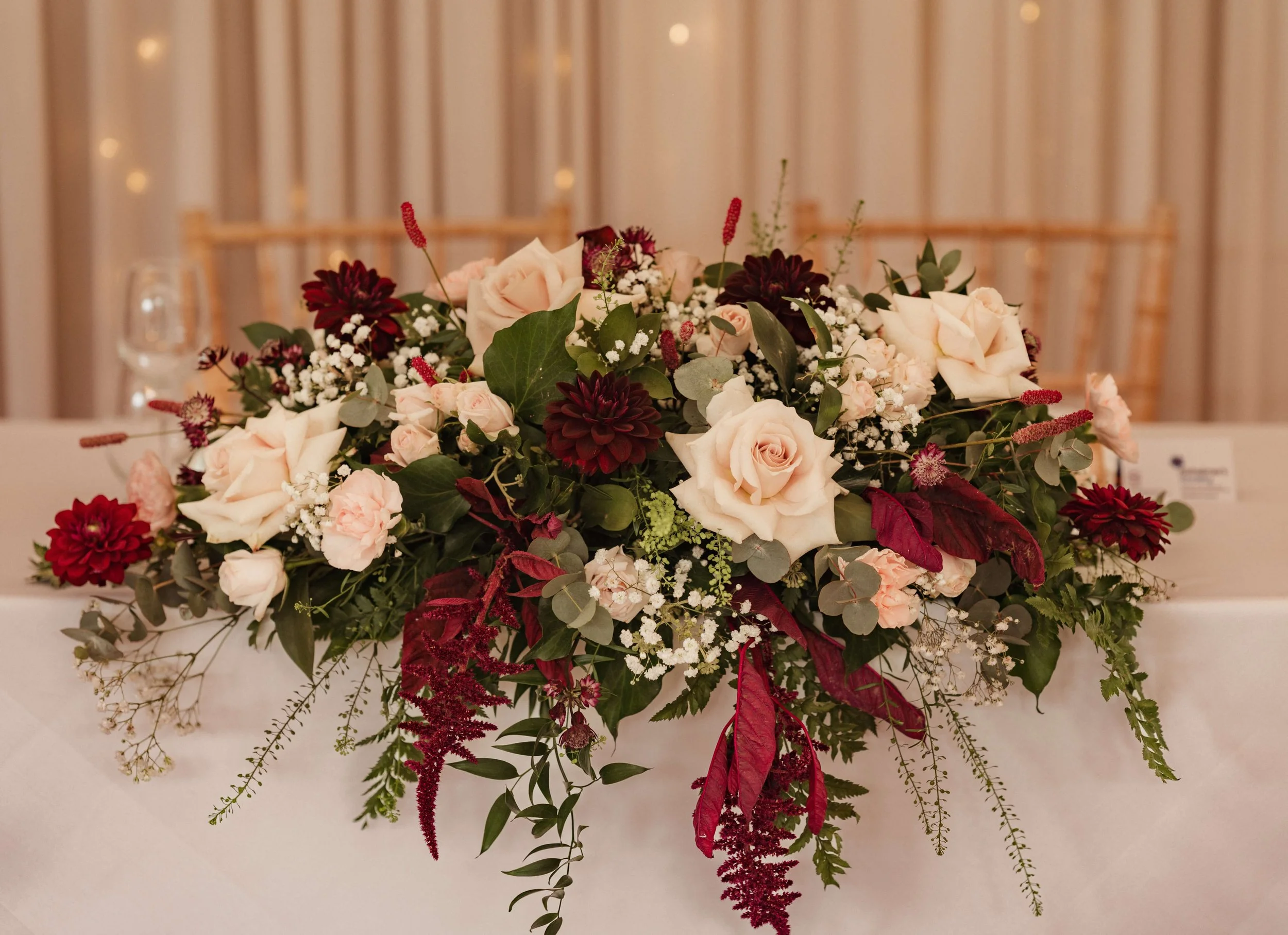 A floral arrangement of light pink roses, dark red dahlias, white baby's breath, and green foliage on a wedding or event table.