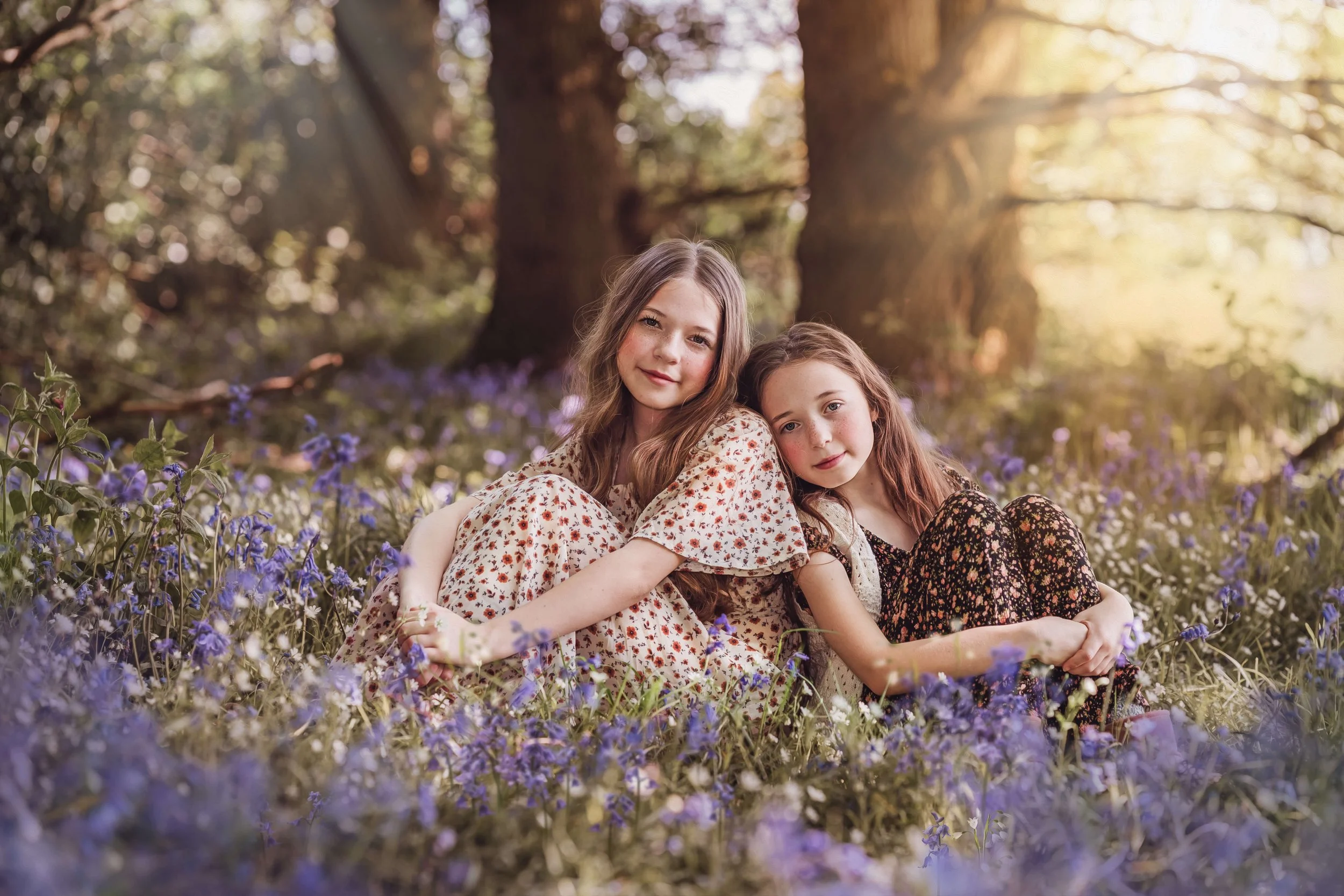 Two young girls sitting in a field of purple wildflowers in a forest, with large trees in the background and warm sunlight filtering through the branches.