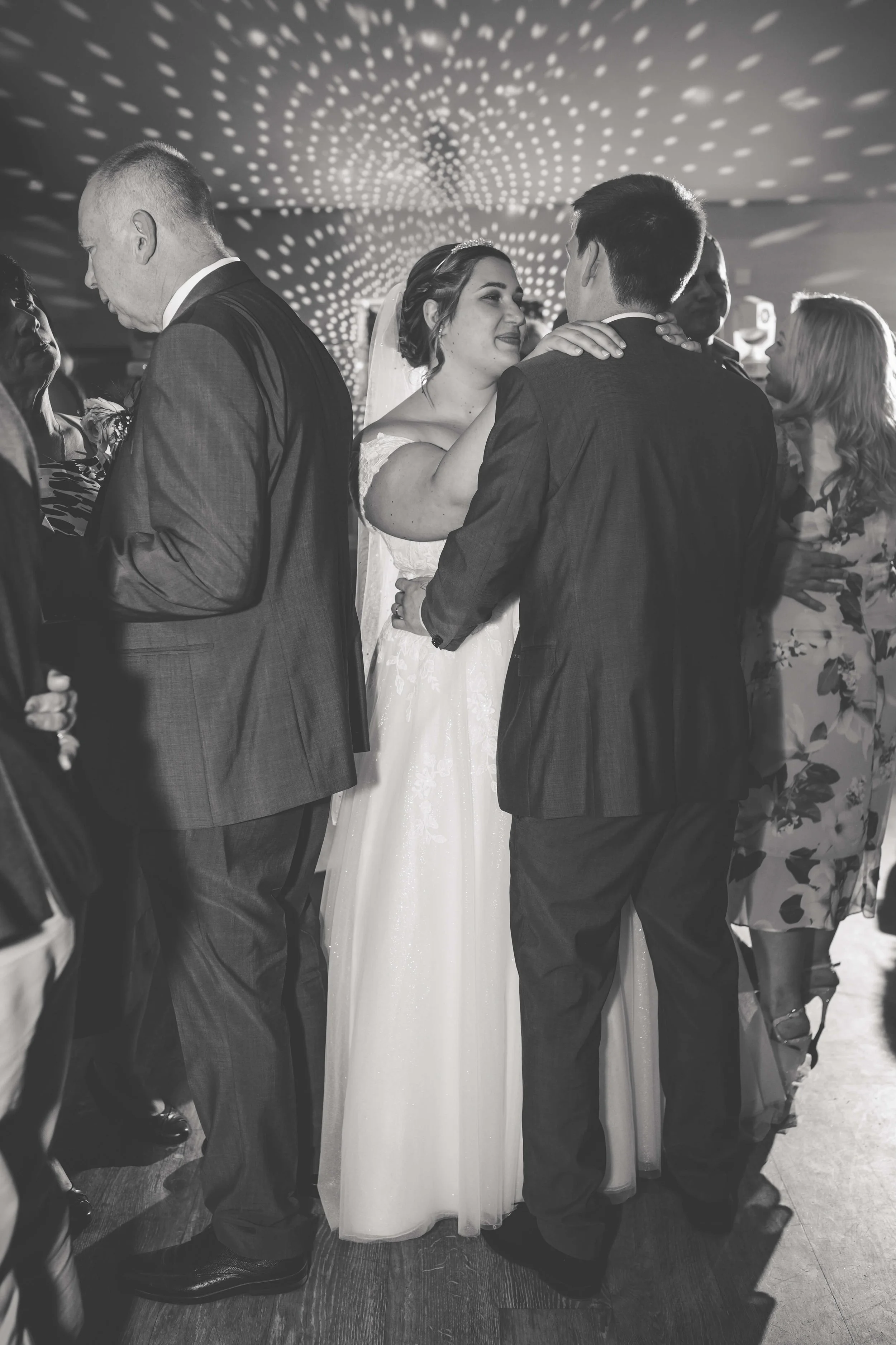 A bride and groom dancing closely at their wedding reception, surrounded by guests under a ceiling light display.