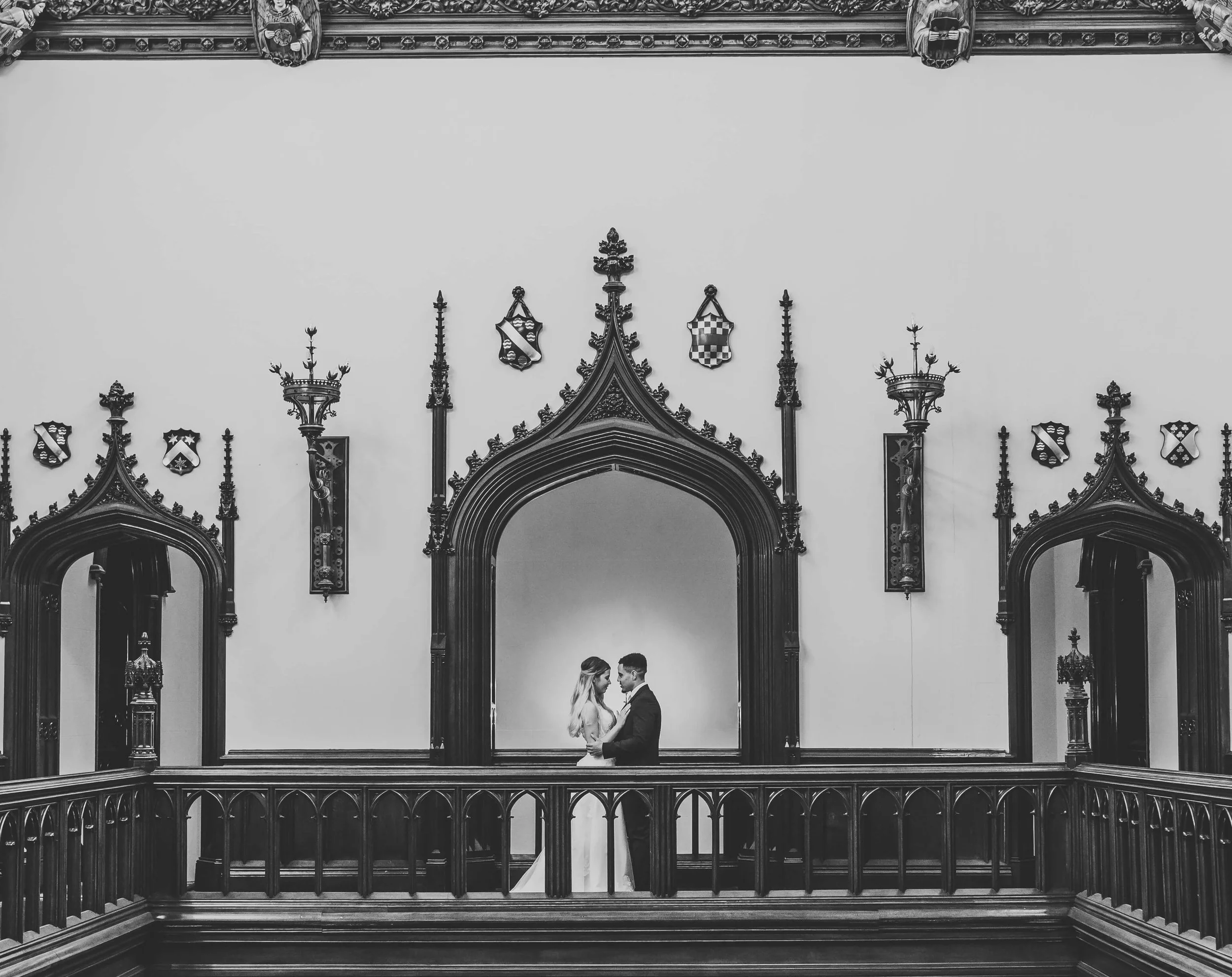 A black and white photograph of a bride and groom standing inside a historic, ornate hall with gothic architectural details, including arched mirrors and decorative shields on the walls. They face each other, close together.