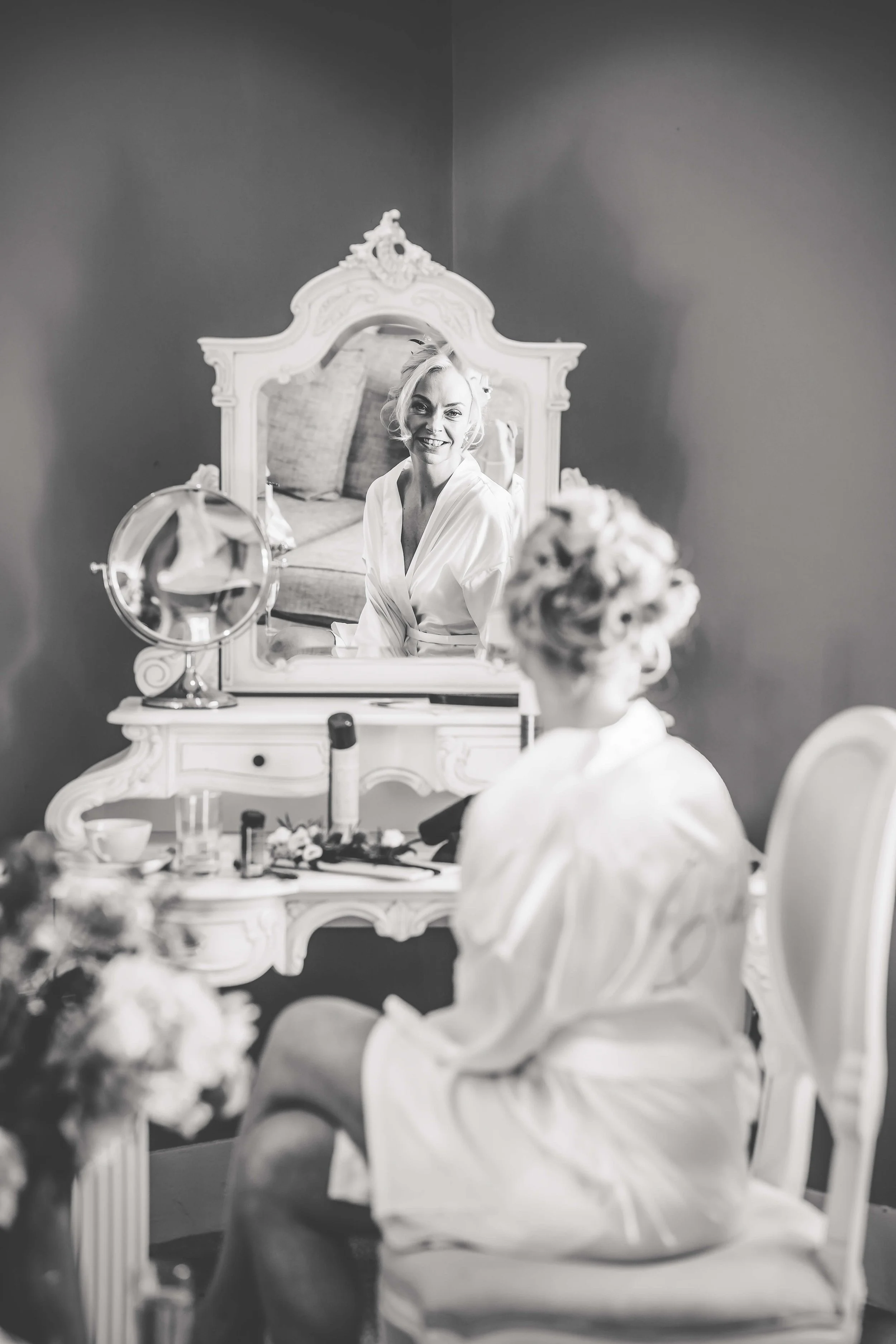 A woman with styled hair and makeup, sitting in front of a mirror, smiling at her reflection, with beauty products on the dresser, in a black and white photo.
