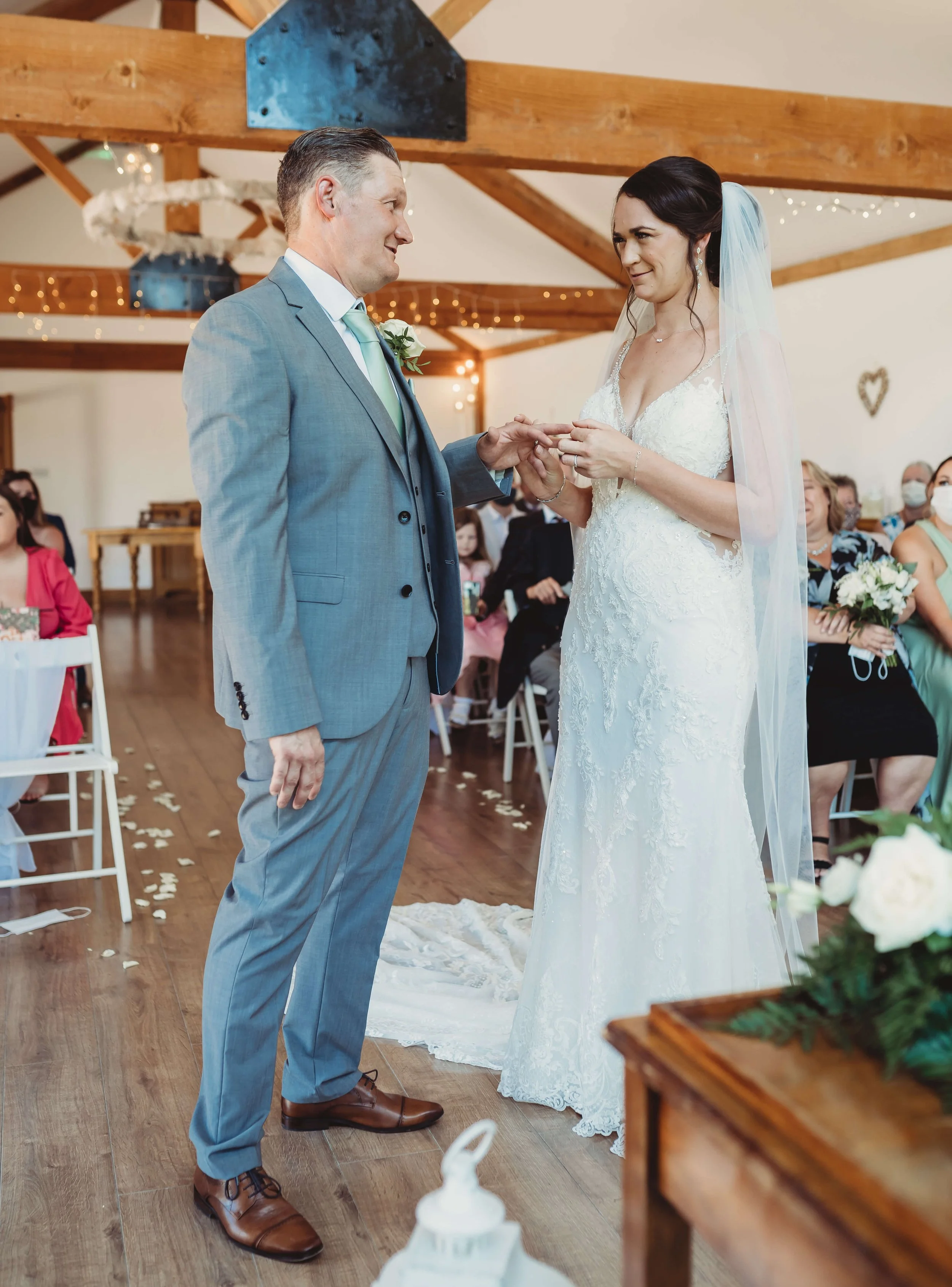 A bride and groom exchange wedding rings during a wedding ceremony in a rustic venue with wooden beams, surrounded by seated guests, some wearing face masks.