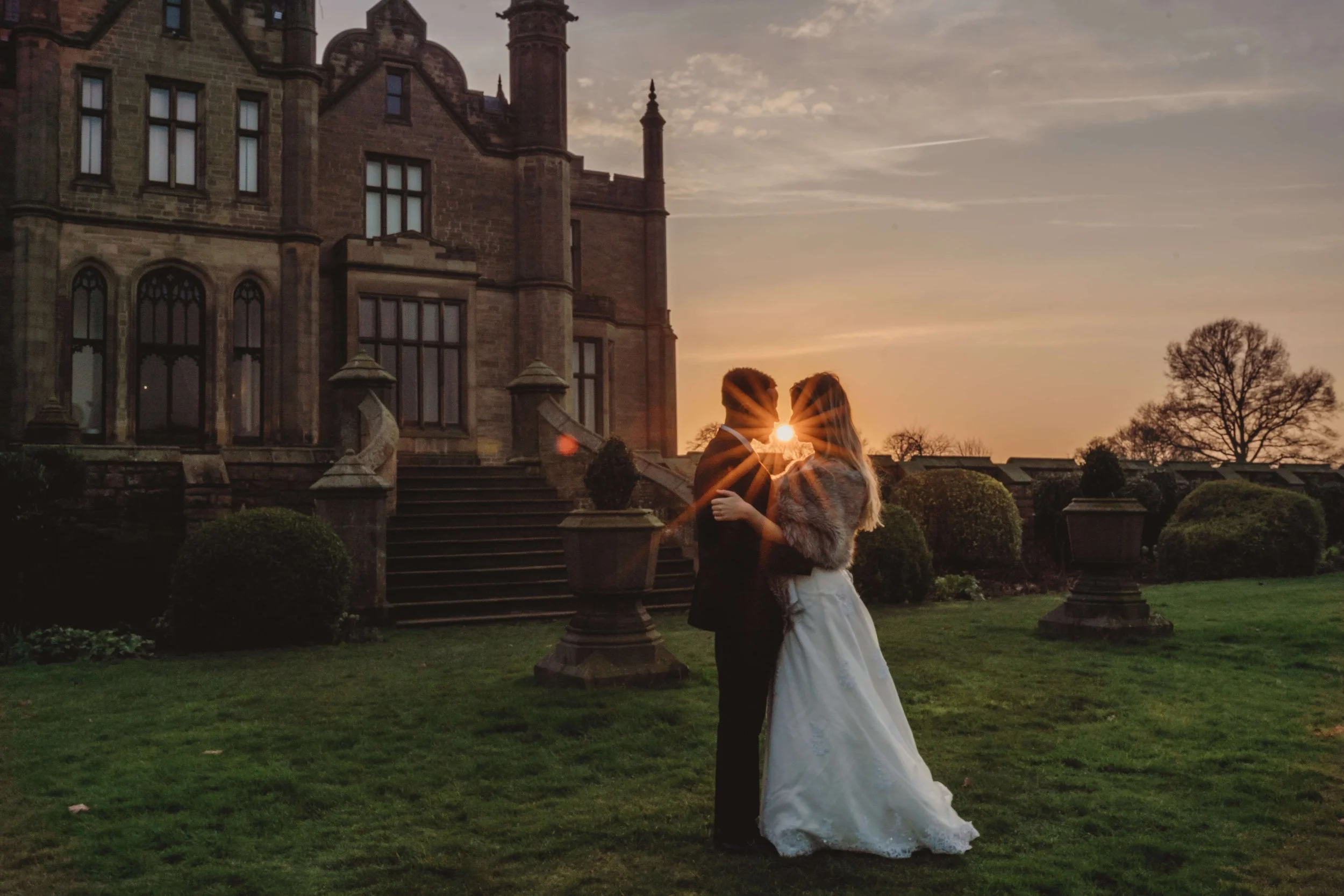A couple in wedding attire stand close together on a lawn at sunset, with the sun positioned between their faces, creating a starburst effect. They are near a historic stone building with stairs and potted plants, and trees are visible in the backgro