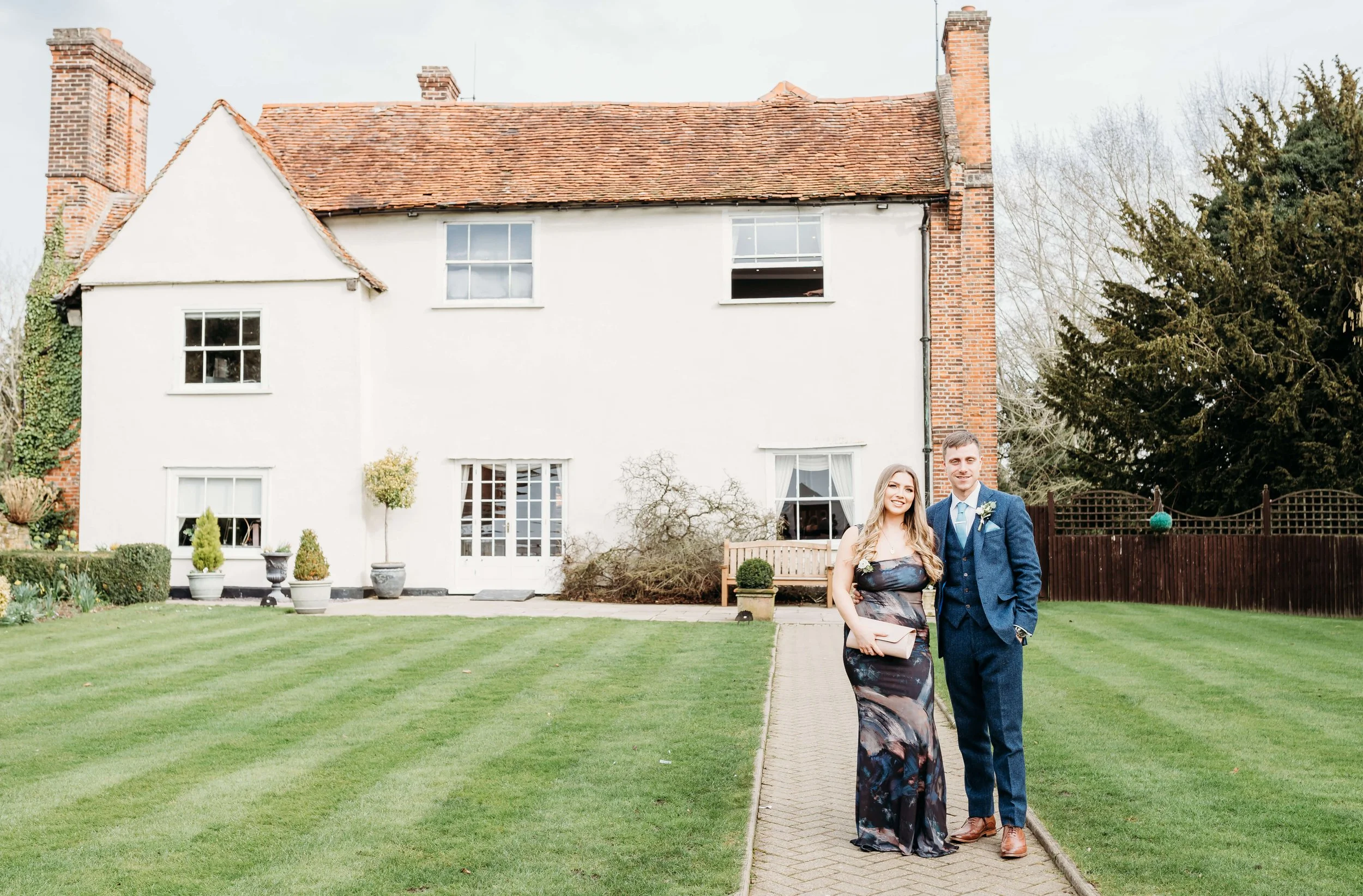 A couple dressed in formal attire standing on a paved pathway in front of a white house with a red tile roof and brick chimneys, surrounded by a well-maintained lawn and gardens.