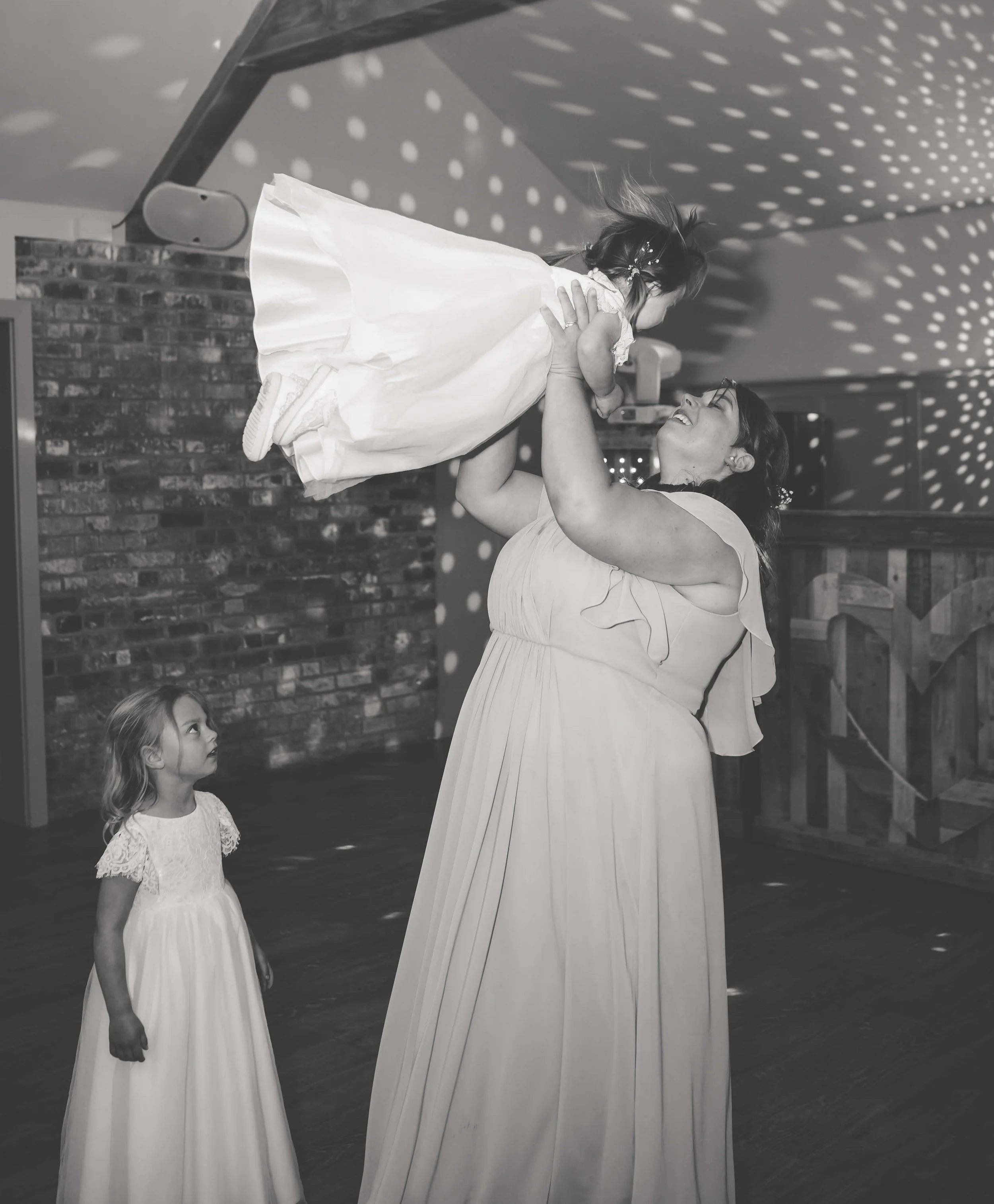 A woman with long dark hair in a light dress lifts a young girl in a white dress, and the girl is looking down at another girl in a white dress, who is watching them.