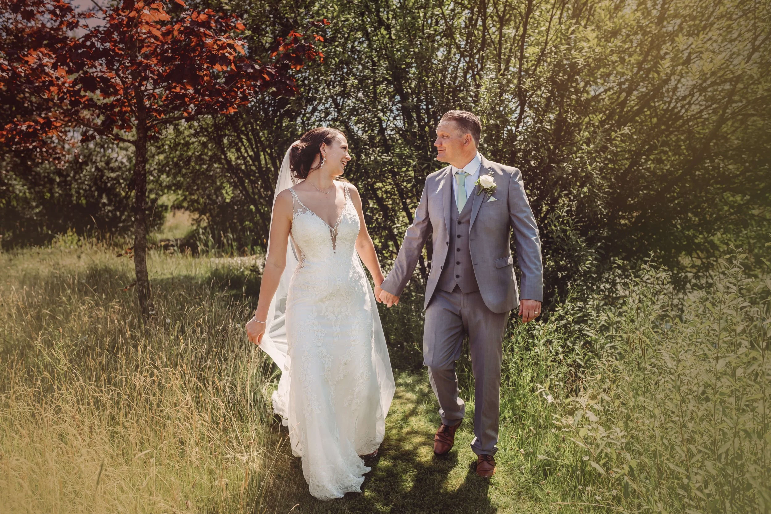 A bride and groom holding hands and smiling at each other as they walk outdoors on a sunny day, surrounded by trees and greenery.