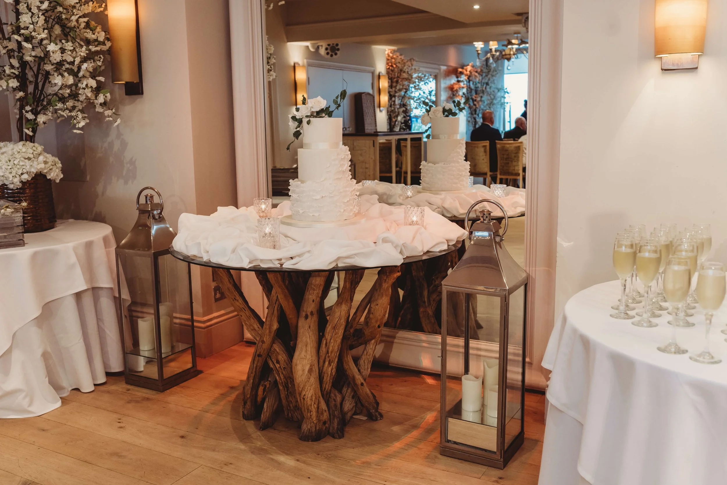 A wedding cake with three tiers topped with white flowers, displayed on a rustic wooden stand, surrounded by candles in glass holders and lanterns, in a decorated reception area.
