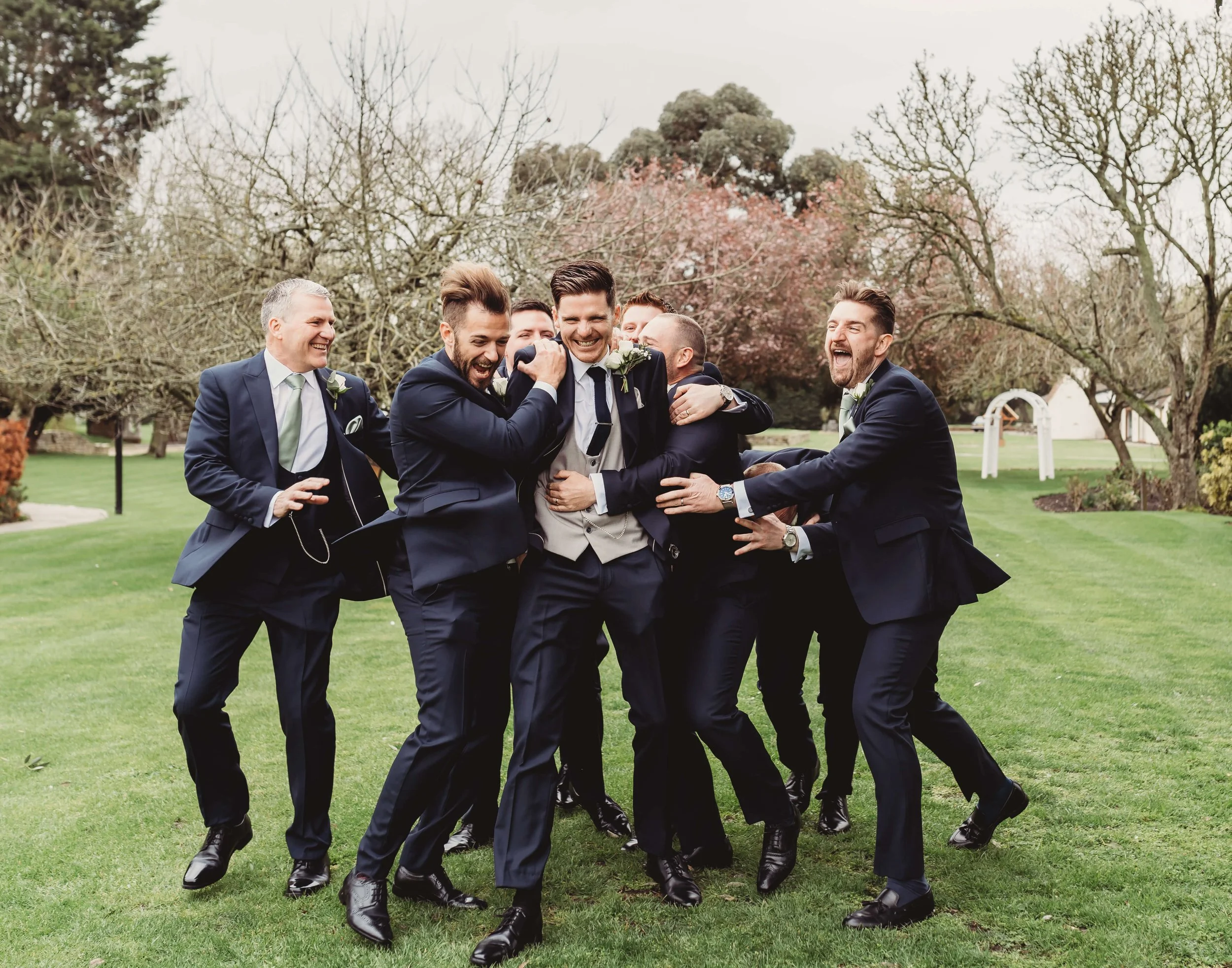 Group of groomsmen in suits laughing and celebrating outdoors on a grassy field with trees and cloudy sky in the background.