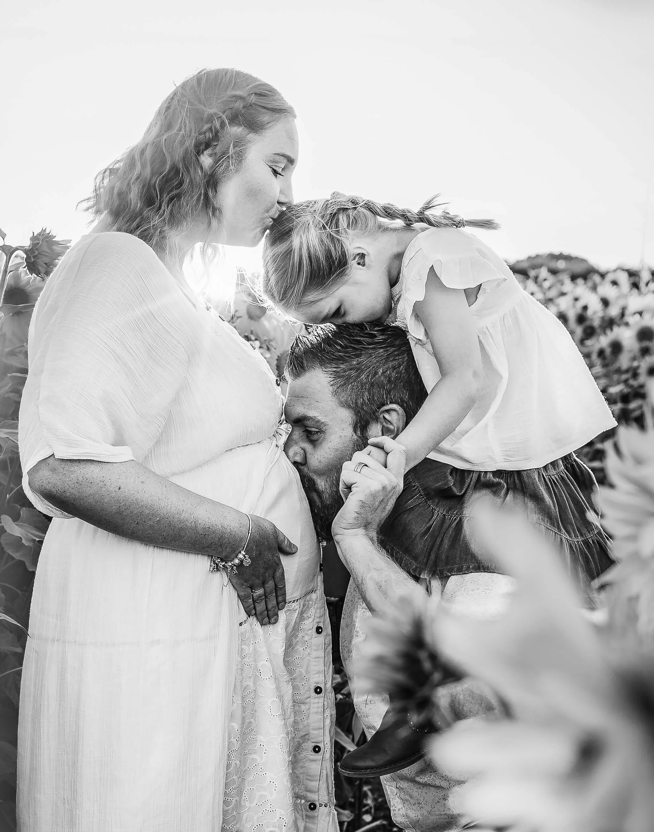 A pregnant woman with a child and a man in a sunflower field. The man is kissing the pregnant woman's belly as the child leans over her, kissing her forehead.