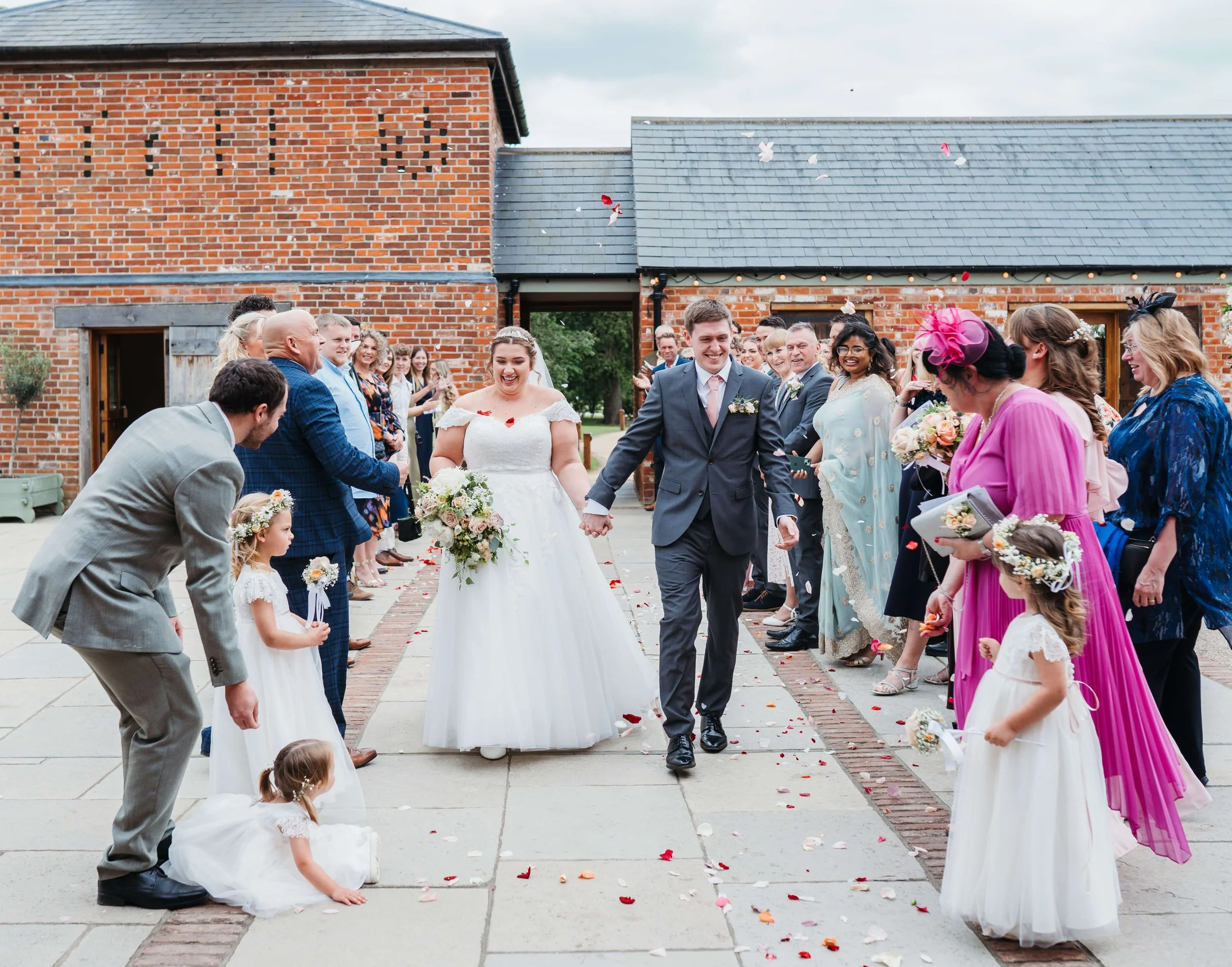 A newlywed couple walks hand in hand down a paved pathway, smiling, while wedding guests on either side throw flower petals. The bride is in a white wedding dress holding a bouquet, and the groom is in a gray suit. Children and adults in formal attir
