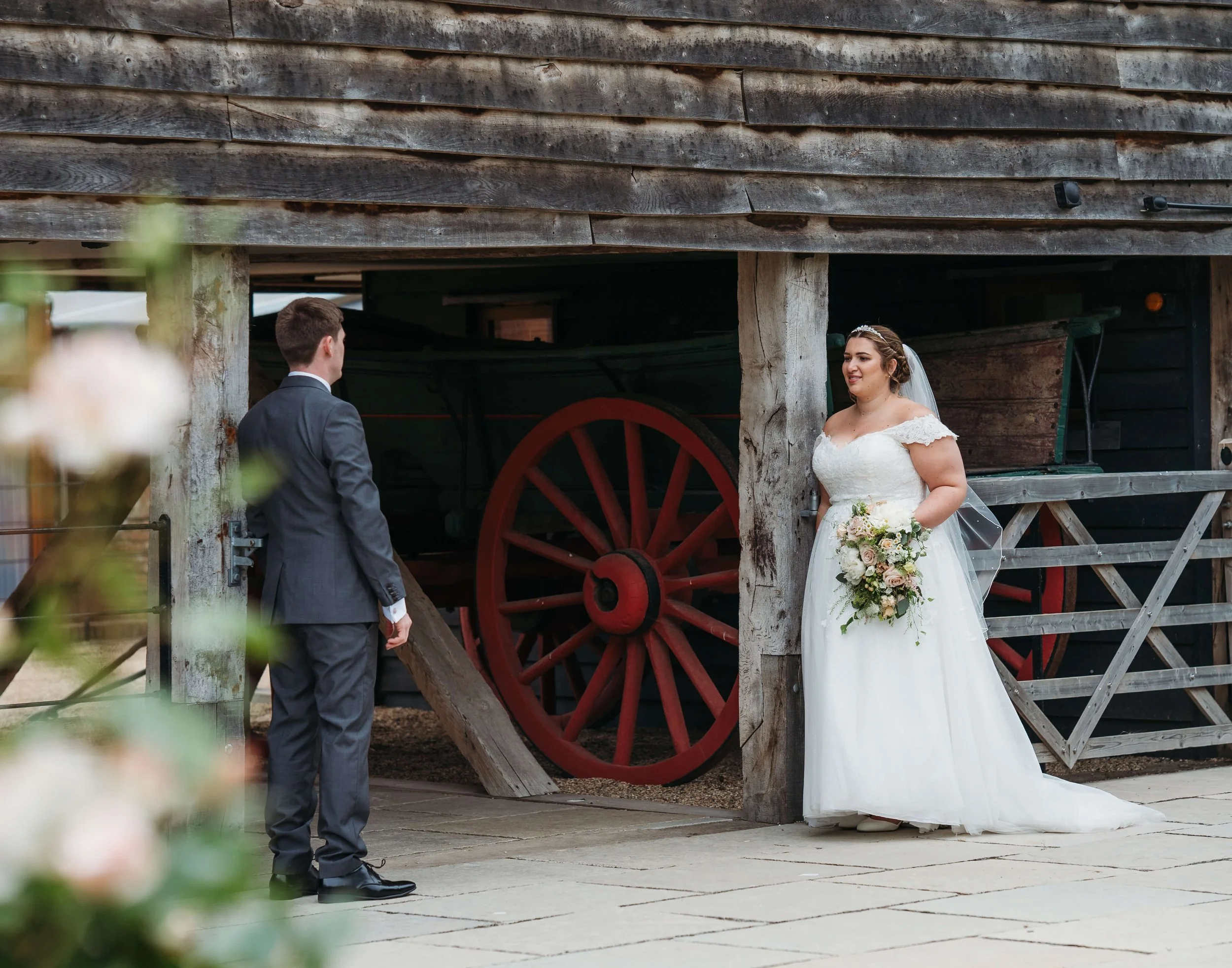 A bride in a white wedding gown holding a bouquet and a groom in a suit facing each other outside a rustic wooden barn with a large red wagon wheel in the background.