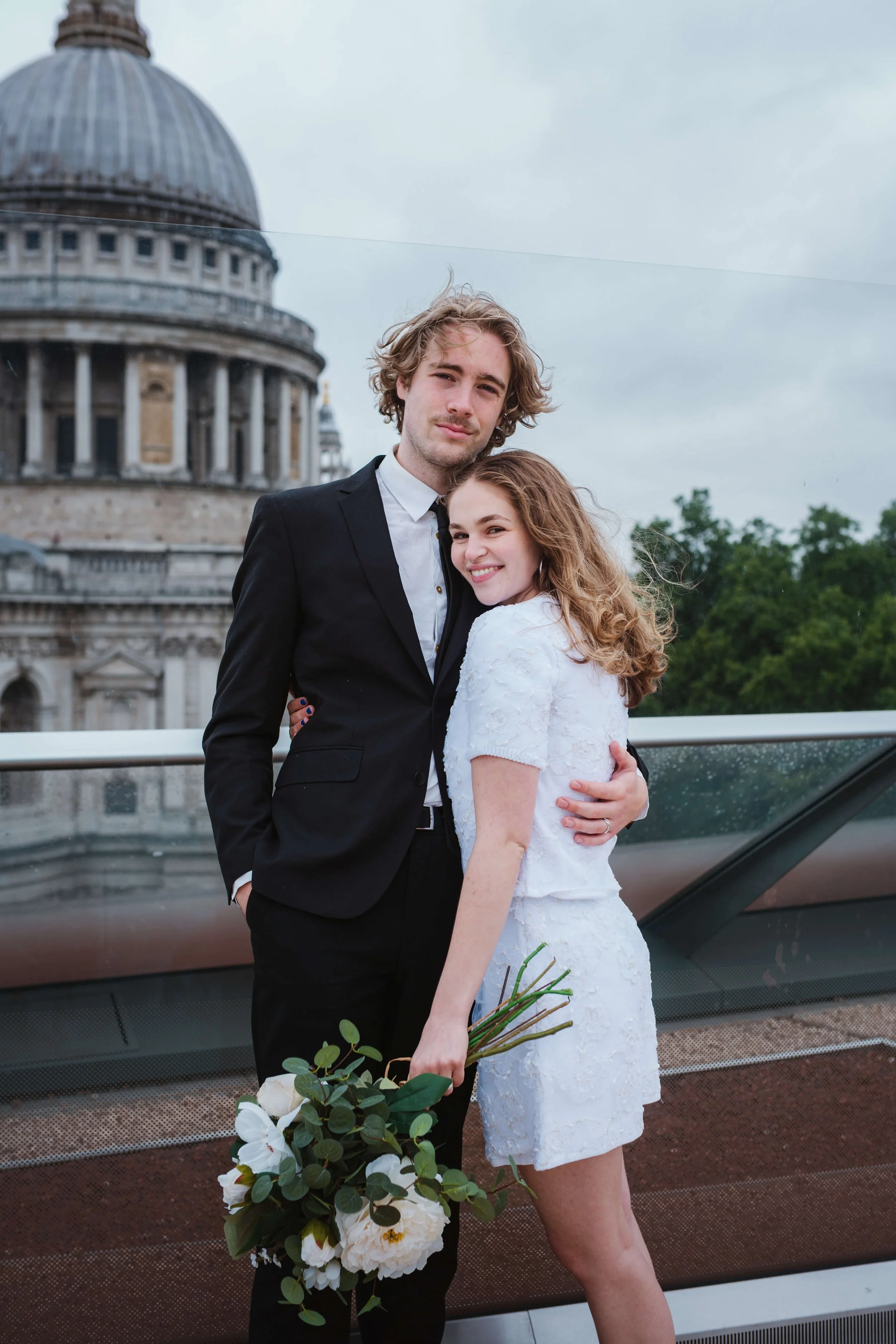 A young couple dressed in formal attire, standing close together outdoors with a historic building with a dome in the background. The woman is holding a bouquet of white flowers and smiling, while the man has his arm around her and is looking at the 