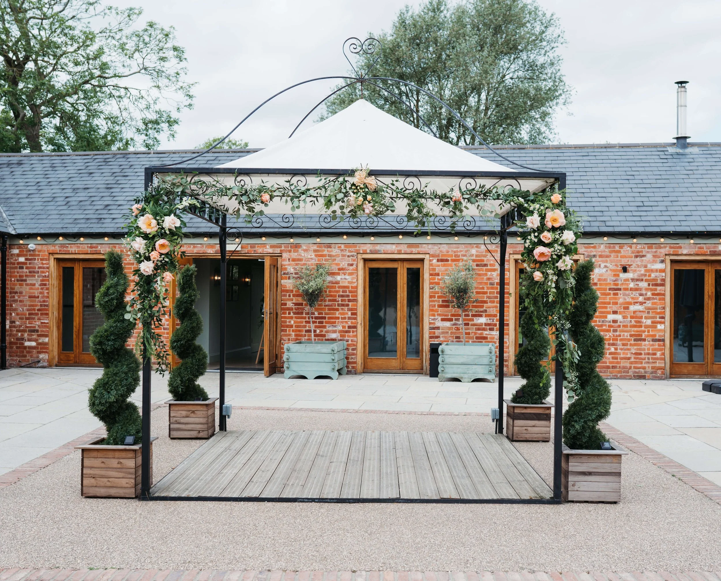 Outdoor wedding or event setup with a decorated black metal arch, pink and white flowers, greenery, and potted plants in front of a brick building with wooden doors.