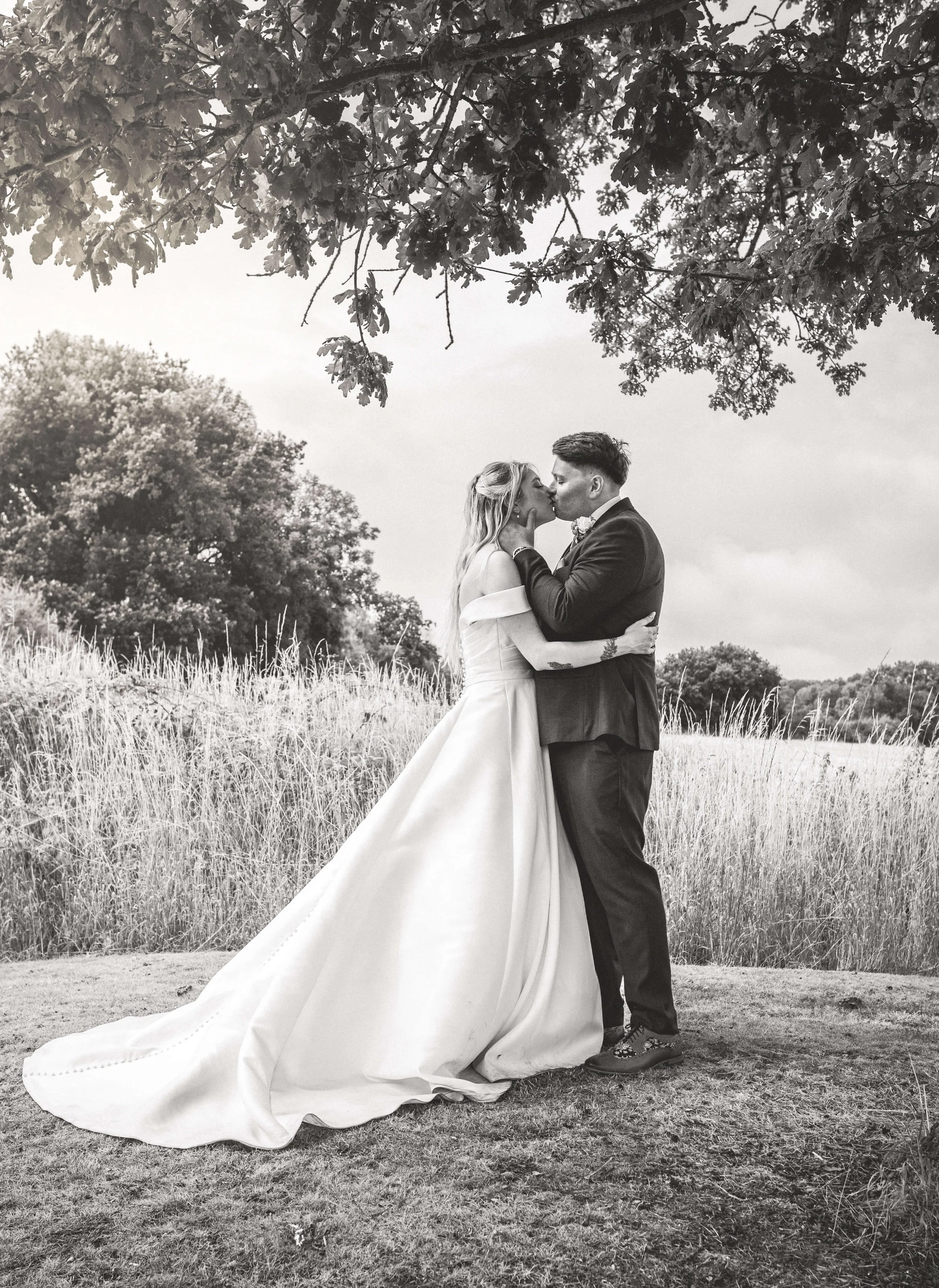 Black and white photo of a bride and groom kissing outdoors, under a tree, with tall grasses and trees in the background.
