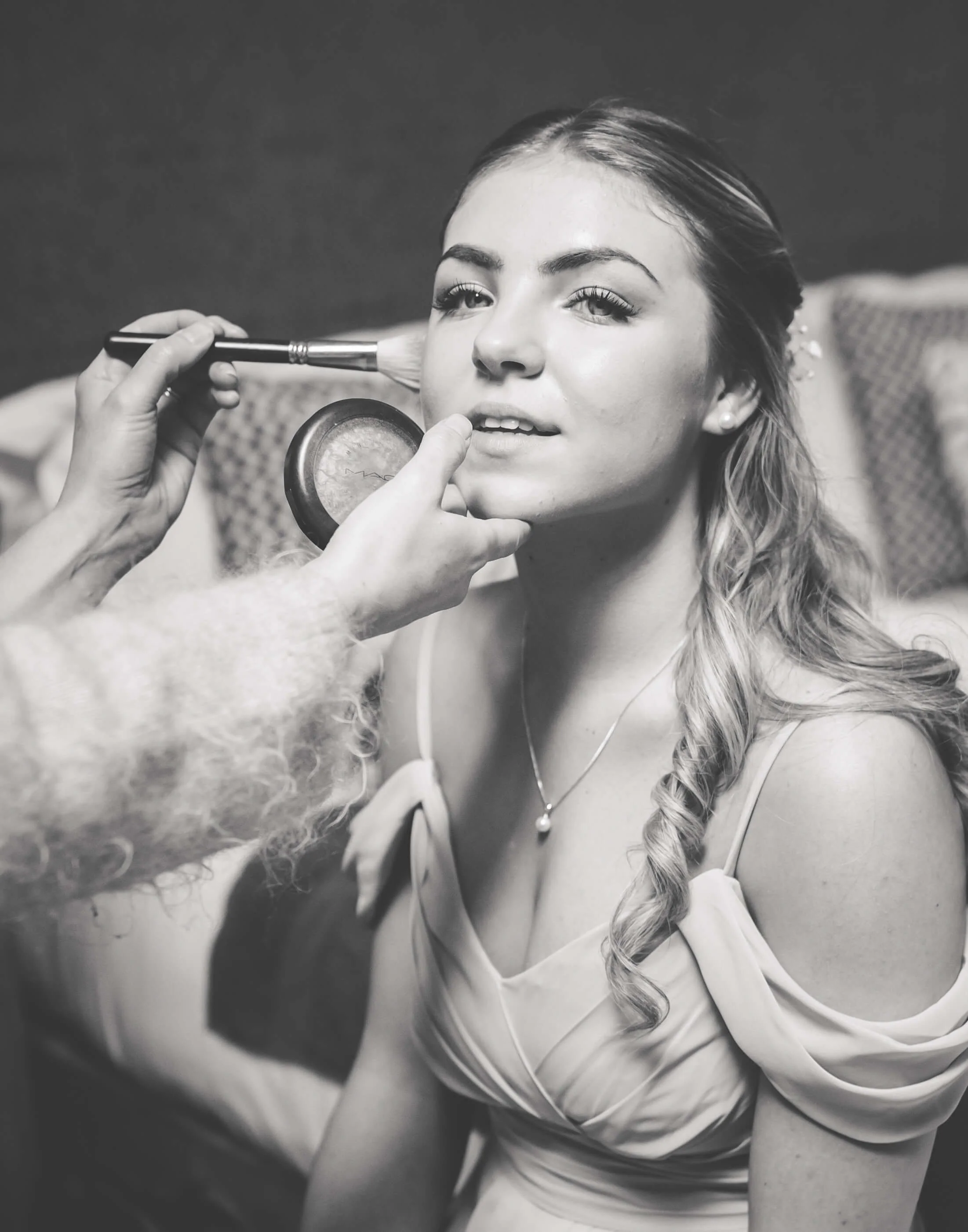 A woman getting her makeup done, with a makeup brush near her face and a makeup compact held by a stylist. The woman has long, curled hair and is wearing a dress with open shoulders and a necklace.