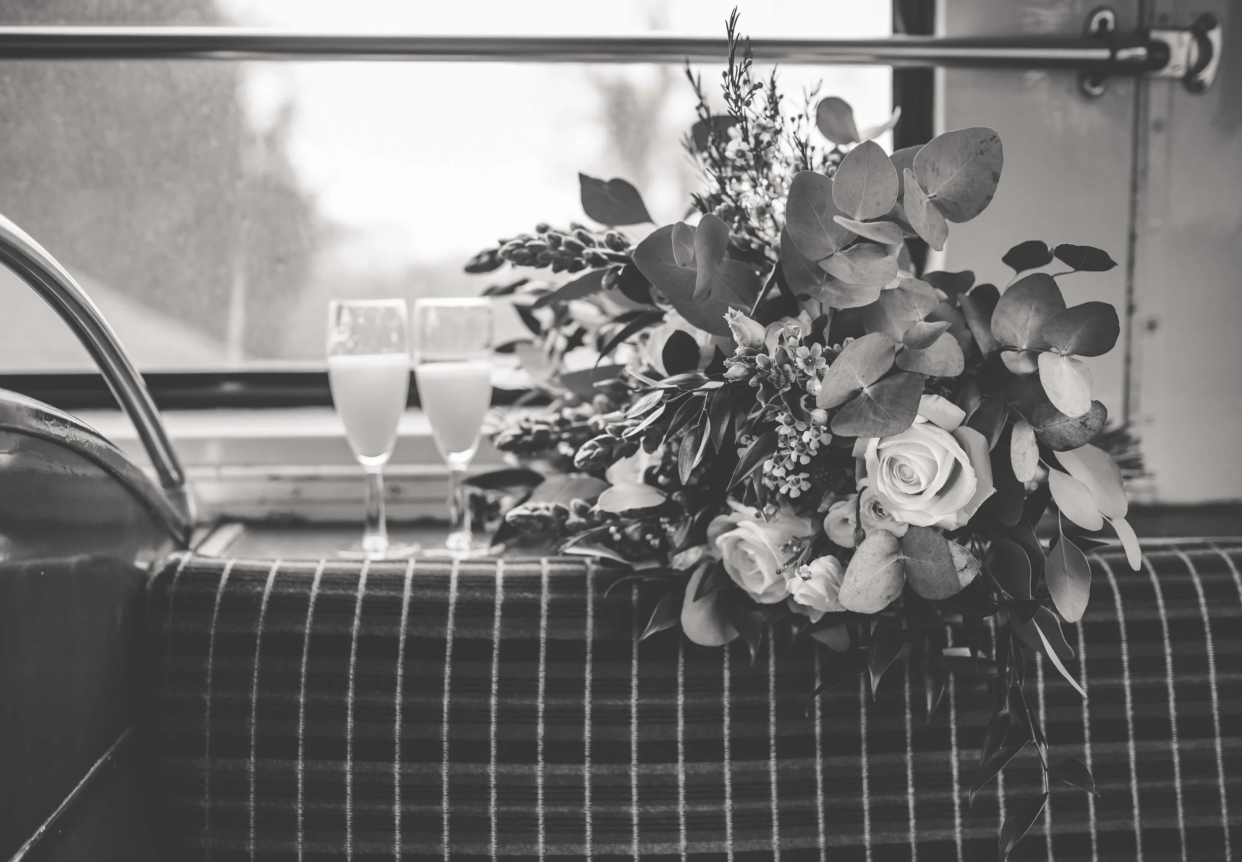 Black and white photo of wedding bouquet with roses and greenery, two flutes of champagne, on a checkered tablecloth by a window.